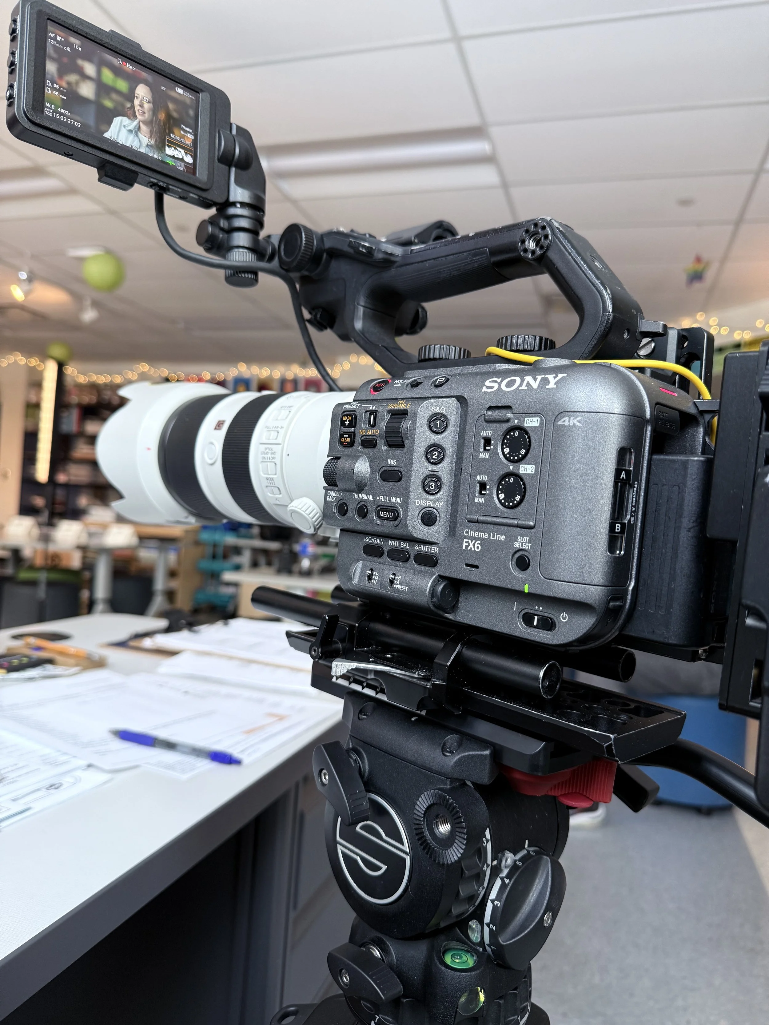 A professional video camera mounted on a tripod, capturing a scene in a studio or workspace with cluttered desks, papers, and pens in the background.