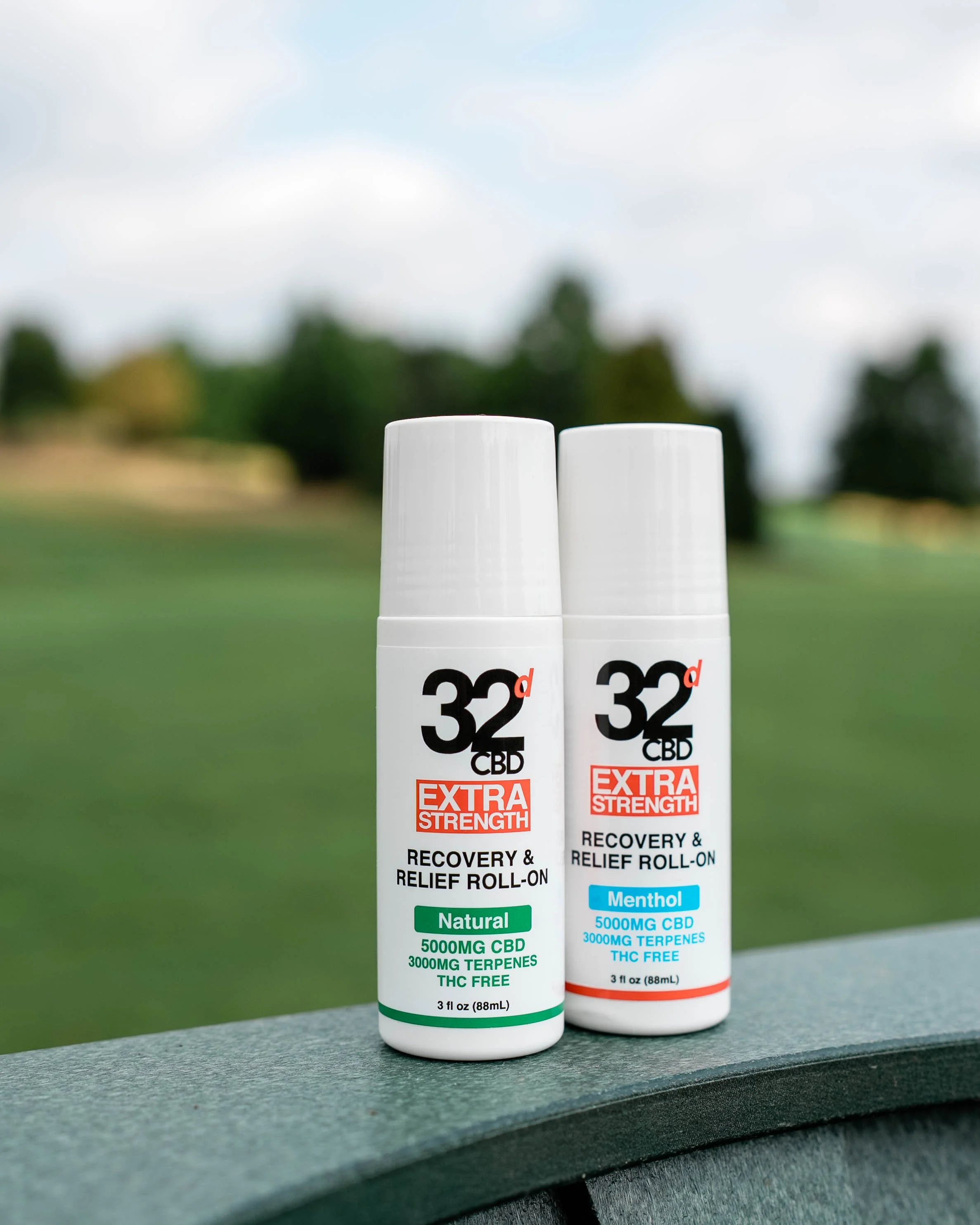 Two bottles of 32d CBD extra strength recovery and relief roll-on, one labeled Natural and the other Menthol, placed outdoors on a bench with a blurred grassy field and trees in the background.