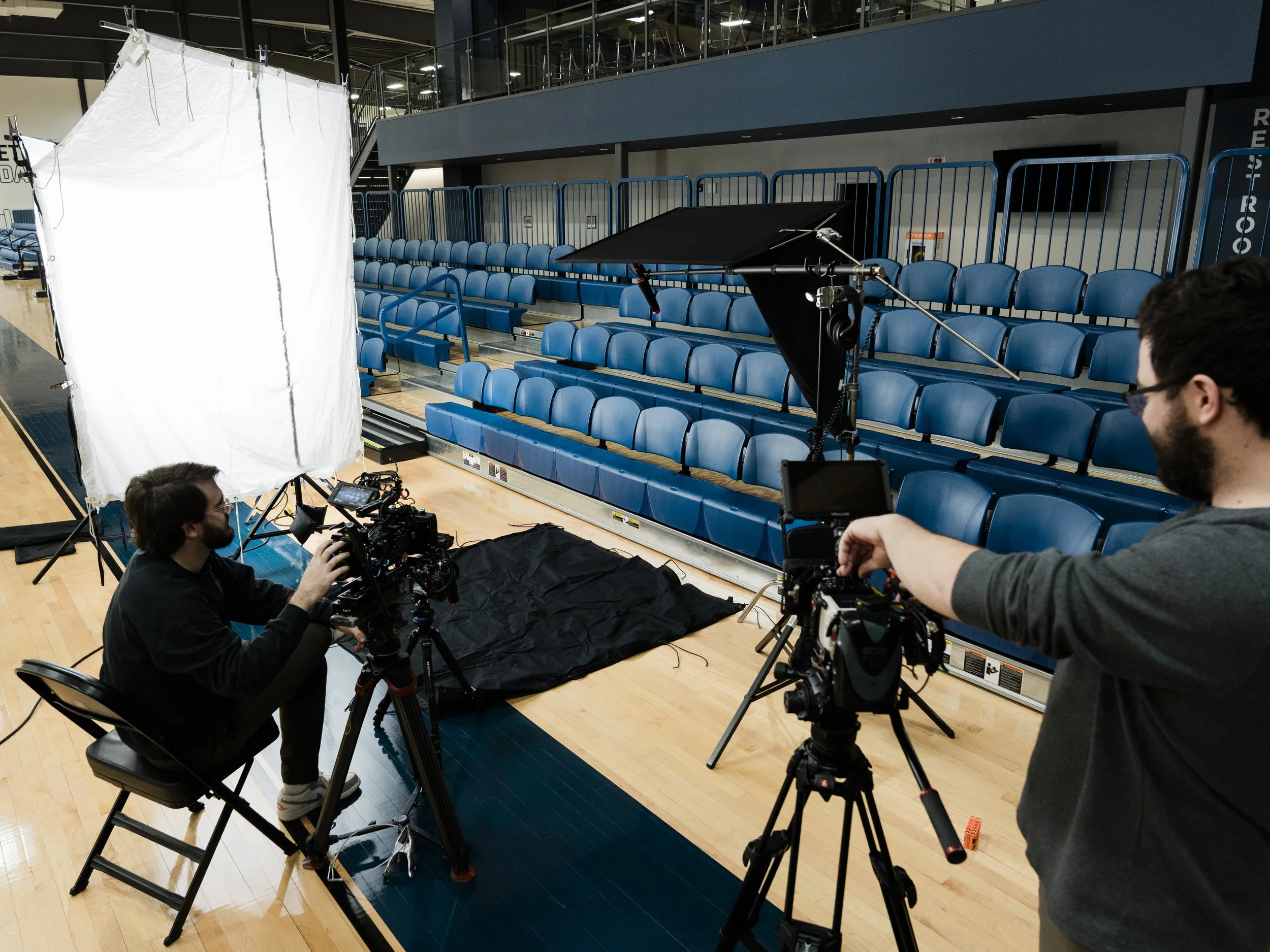 Photography setup in an indoor sports arena with blue bleacher seats, studio lighting, and camera operators preparing for filming.