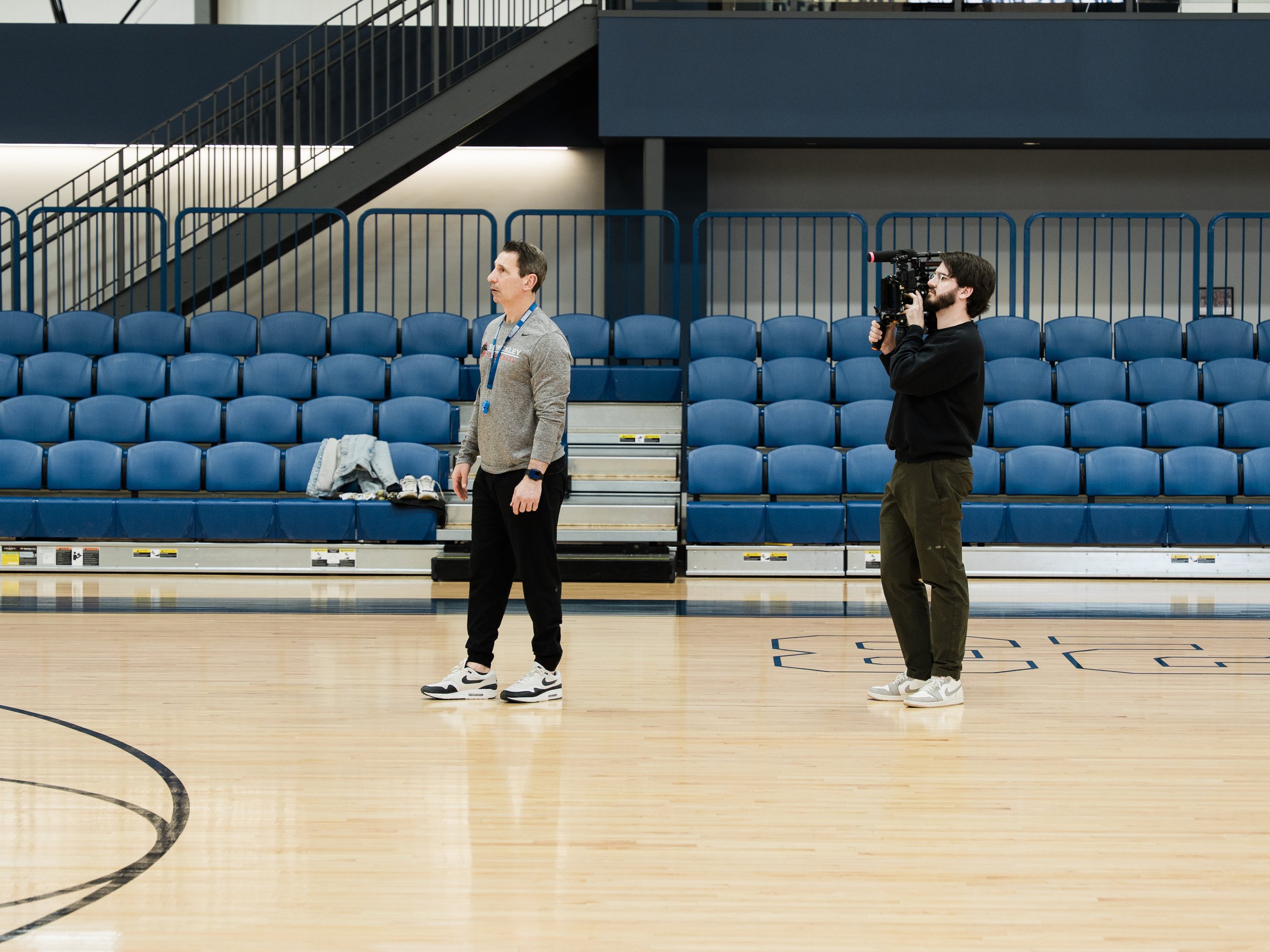 Two men standing on a basketball court inside an arena with blue seating and a stairway in the background. One man is holding a video camera, filming the other man who is standing with his hands by his side, wearing athletic shoes and a training kit.