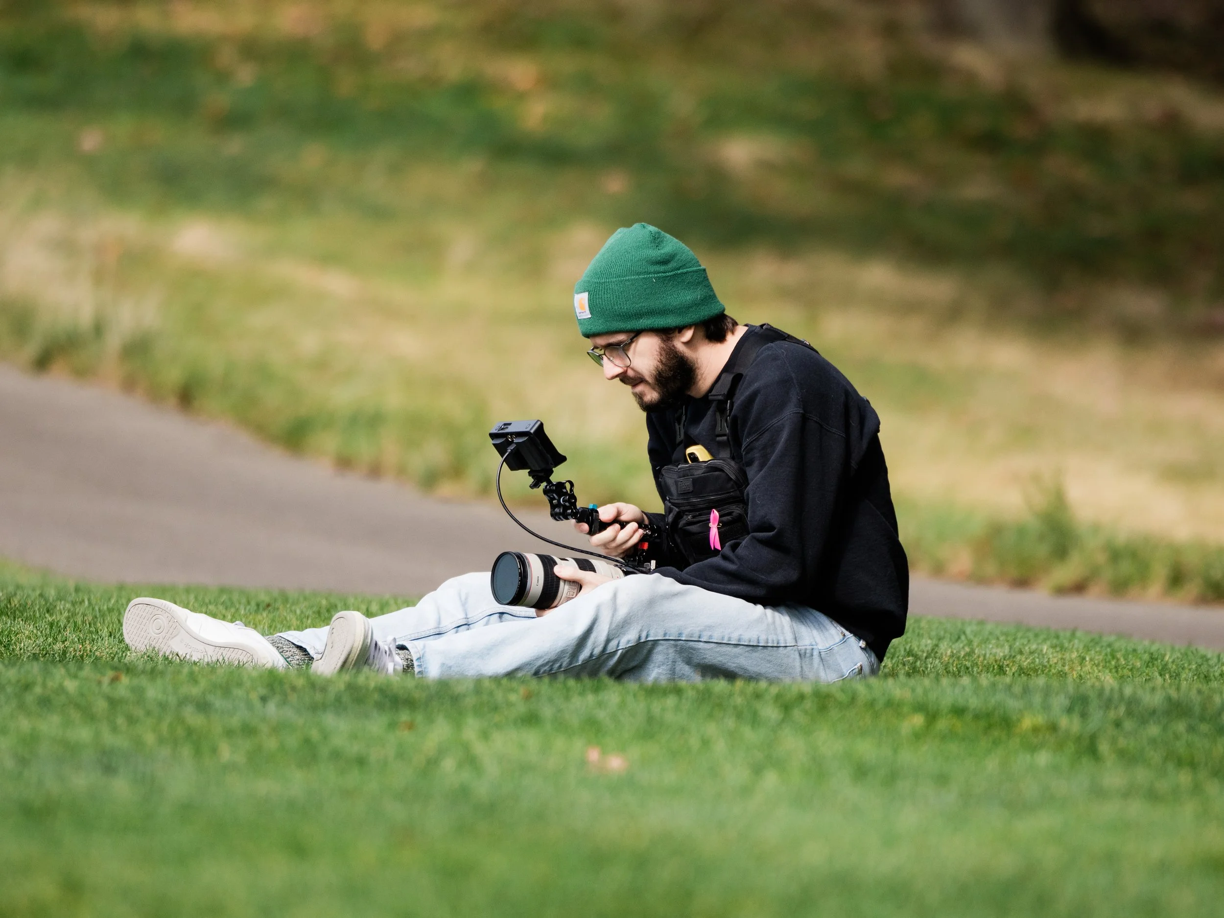 A man with a beard, glasses, and a green beanie wearing a black jacket, sitting on the grass, looking at a camera mounted on a stabilizer, with another person lying on the grass with their legs extended in front of him, in a park or outdoor setting.