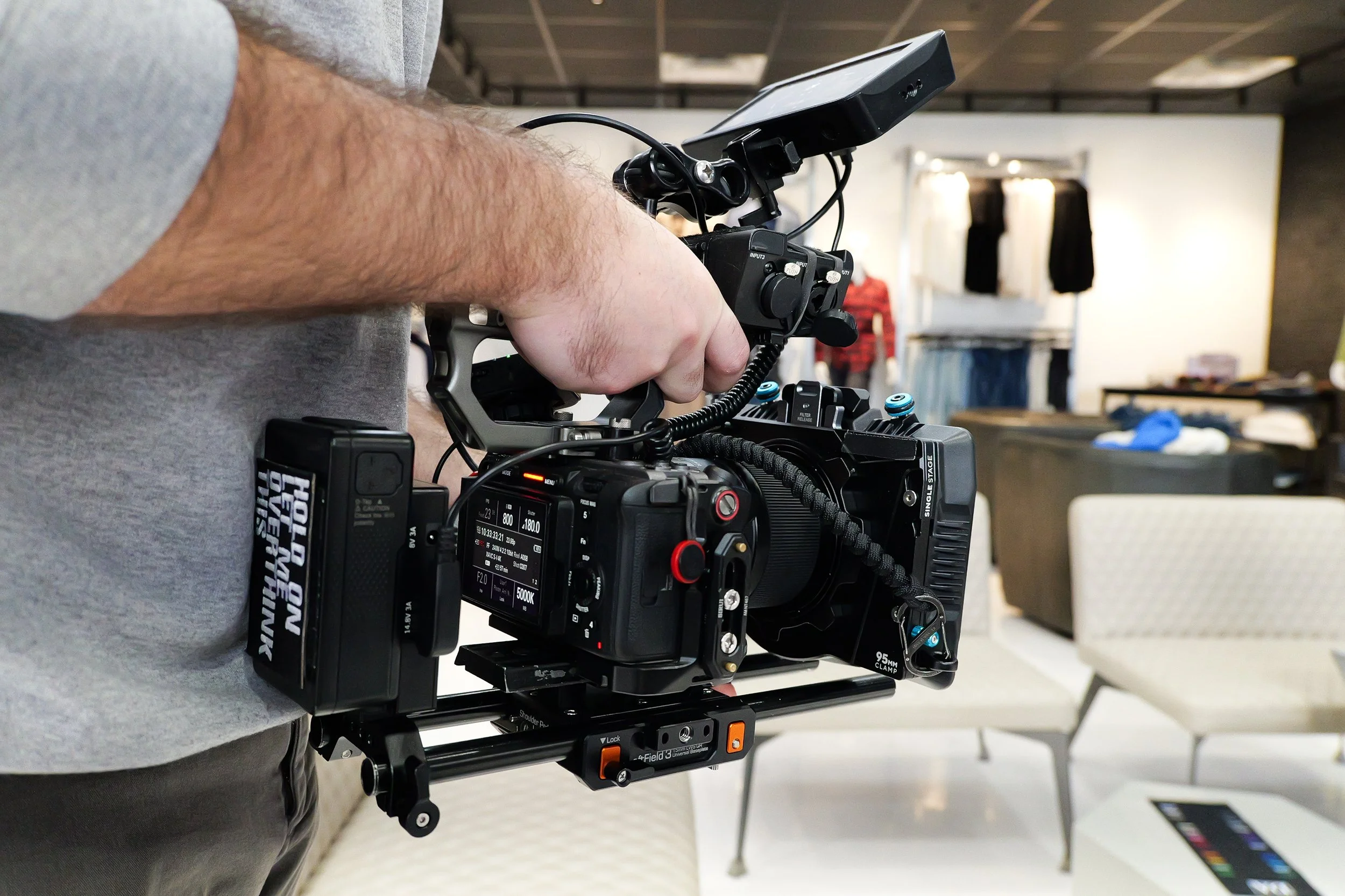 A person holding a professional video camera on a stabilizer, capturing a shot in an indoor setting with furniture and other objects in the background.