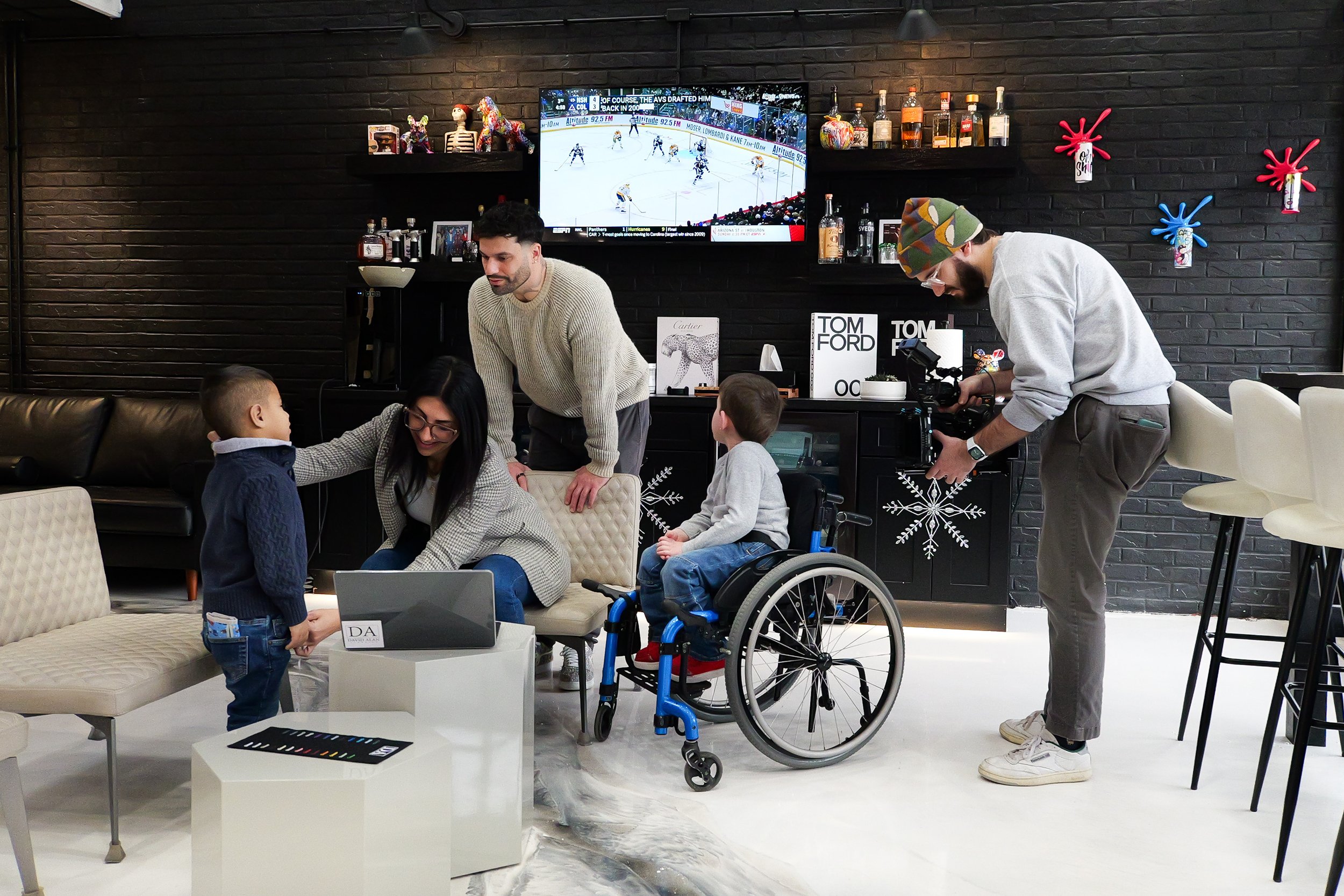 People in a modern indoor space with dark brick walls, watching a hockey game on a large TV screen, and filming a scene with two children, one in a wheelchair. The area has shelves with bottles and art decorations, including colorful animal sculpture