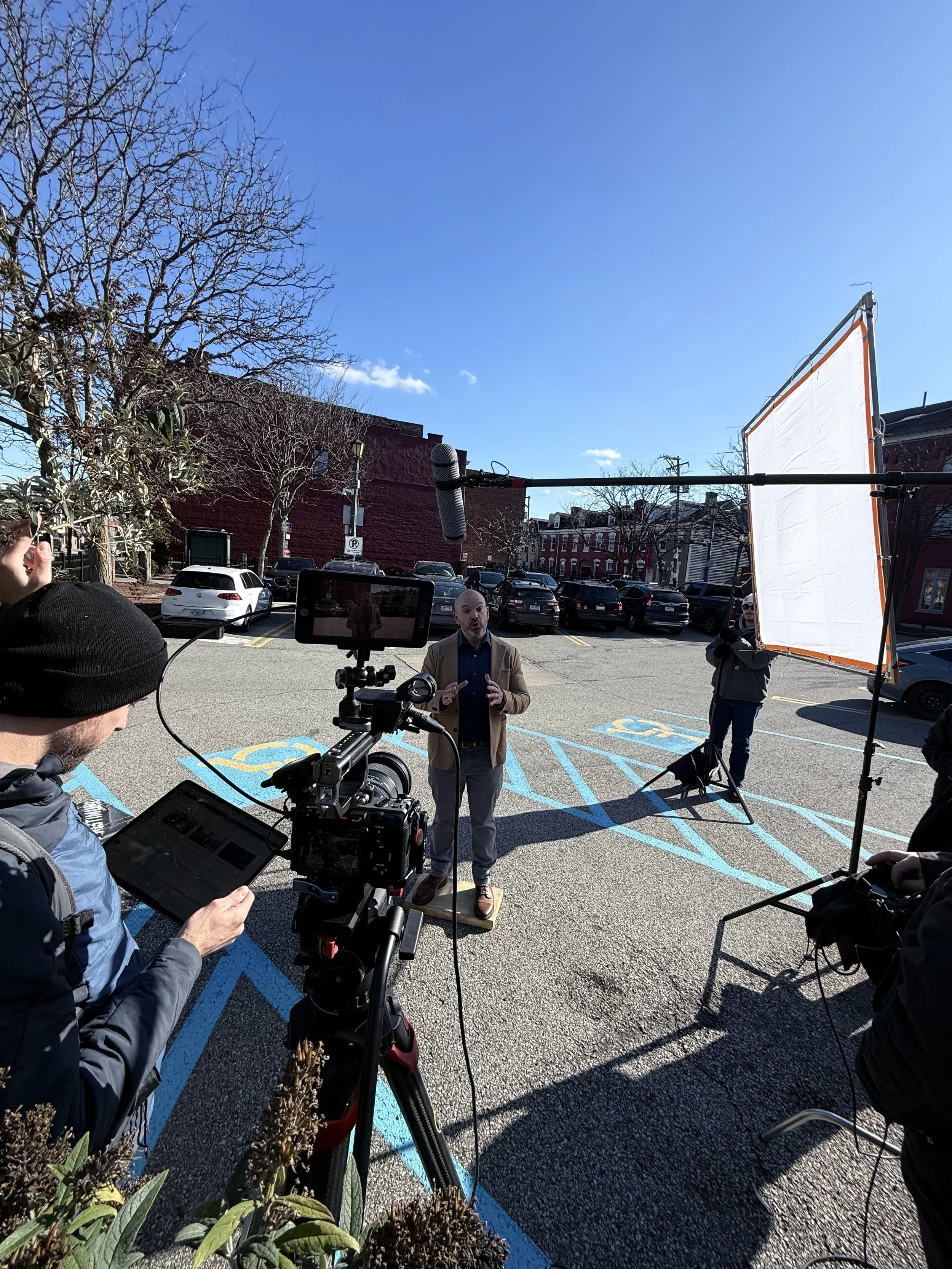 A man in a tan blazer speaks during an outdoor interview or recording session in a parking lot with blue handicapped parking lines, surrounded by screening equipment, a camera, a microphone, and a crew member holding a clipboard. The background shows