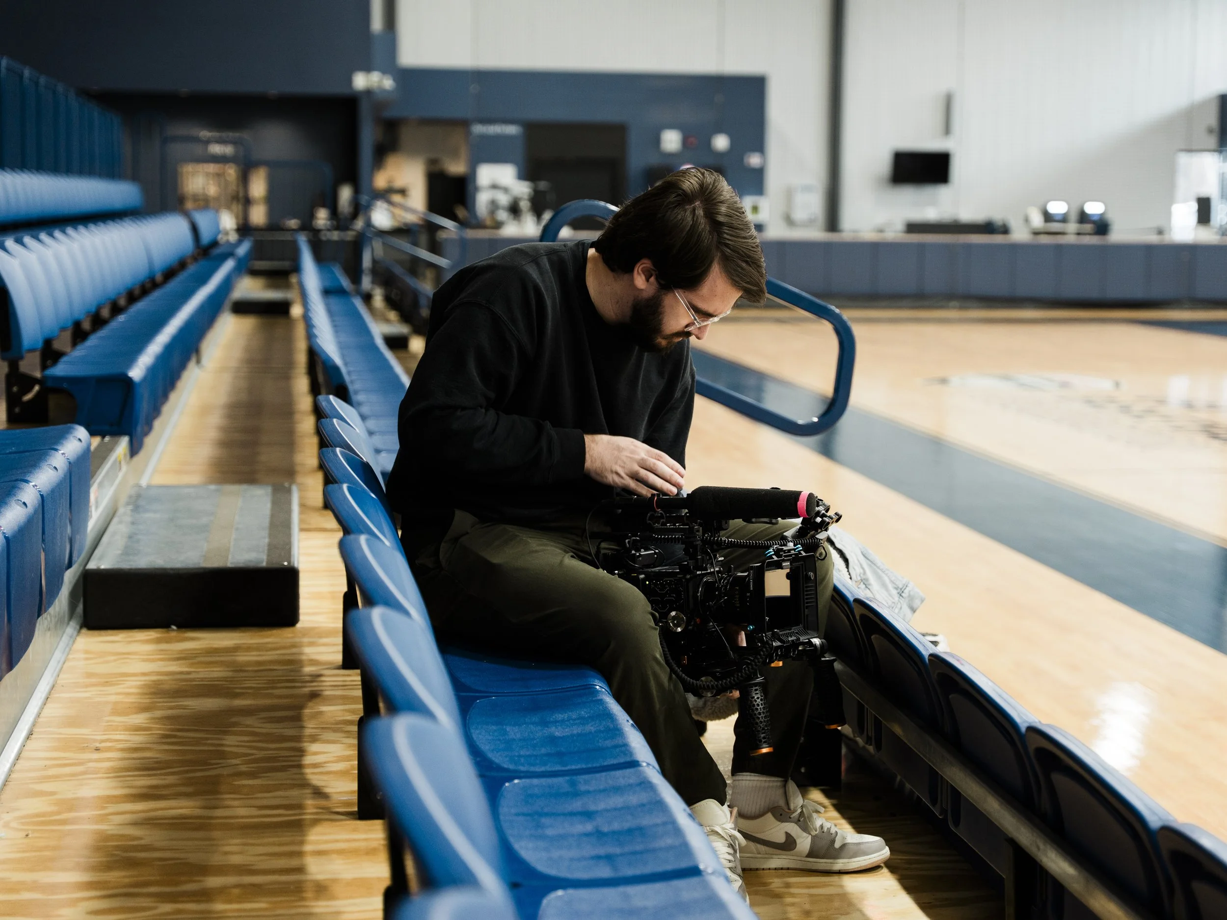 A man with glasses and a beard, sitting alone on blue bleacher seats in an indoor sports arena, appears to be filming or setting up camera equipment.