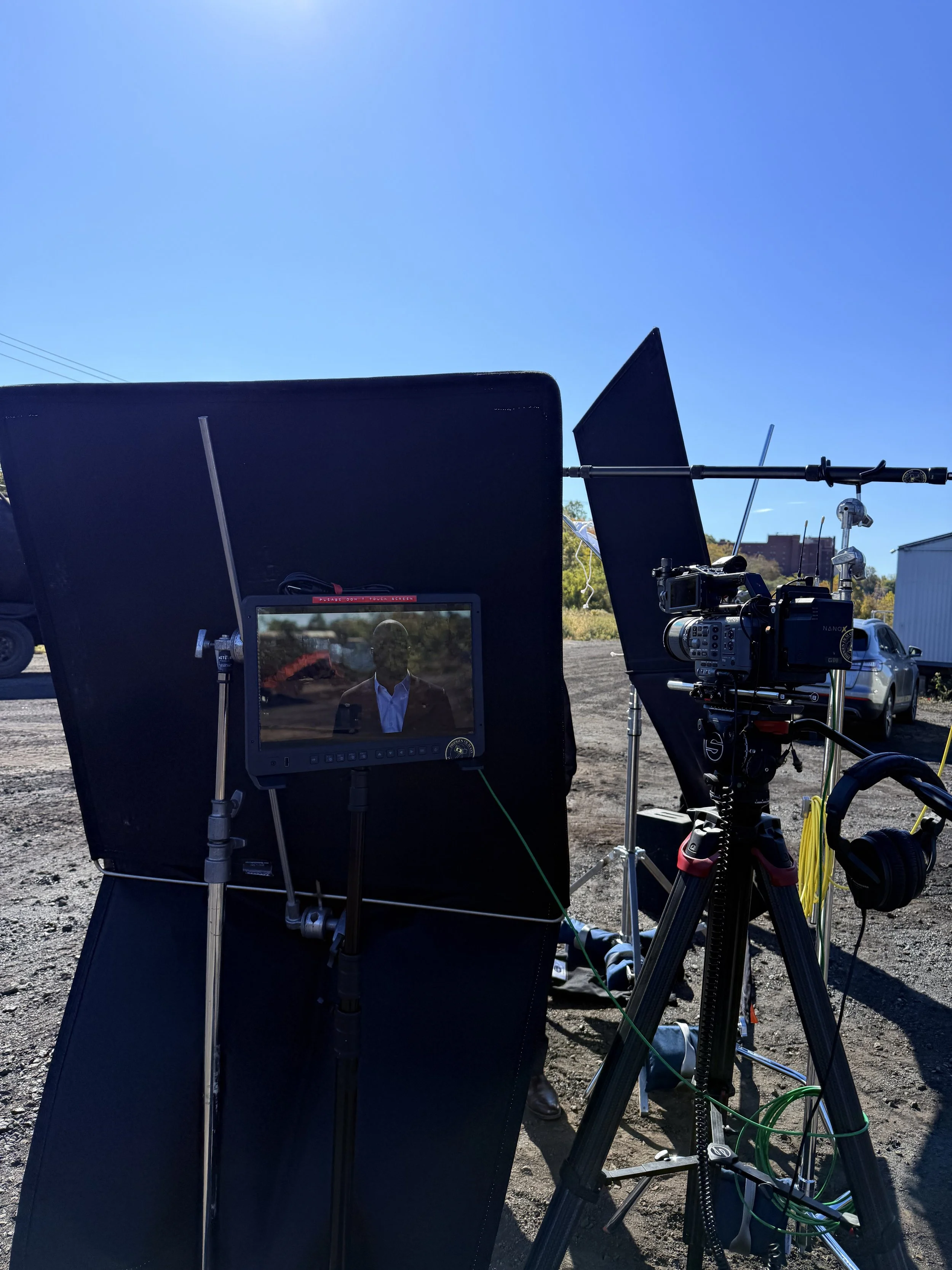 Equipment setup for a film or video shoot outdoors, including a monitor displaying a man in a suit, various lights and stands, and camera gear against a clear blue sky.