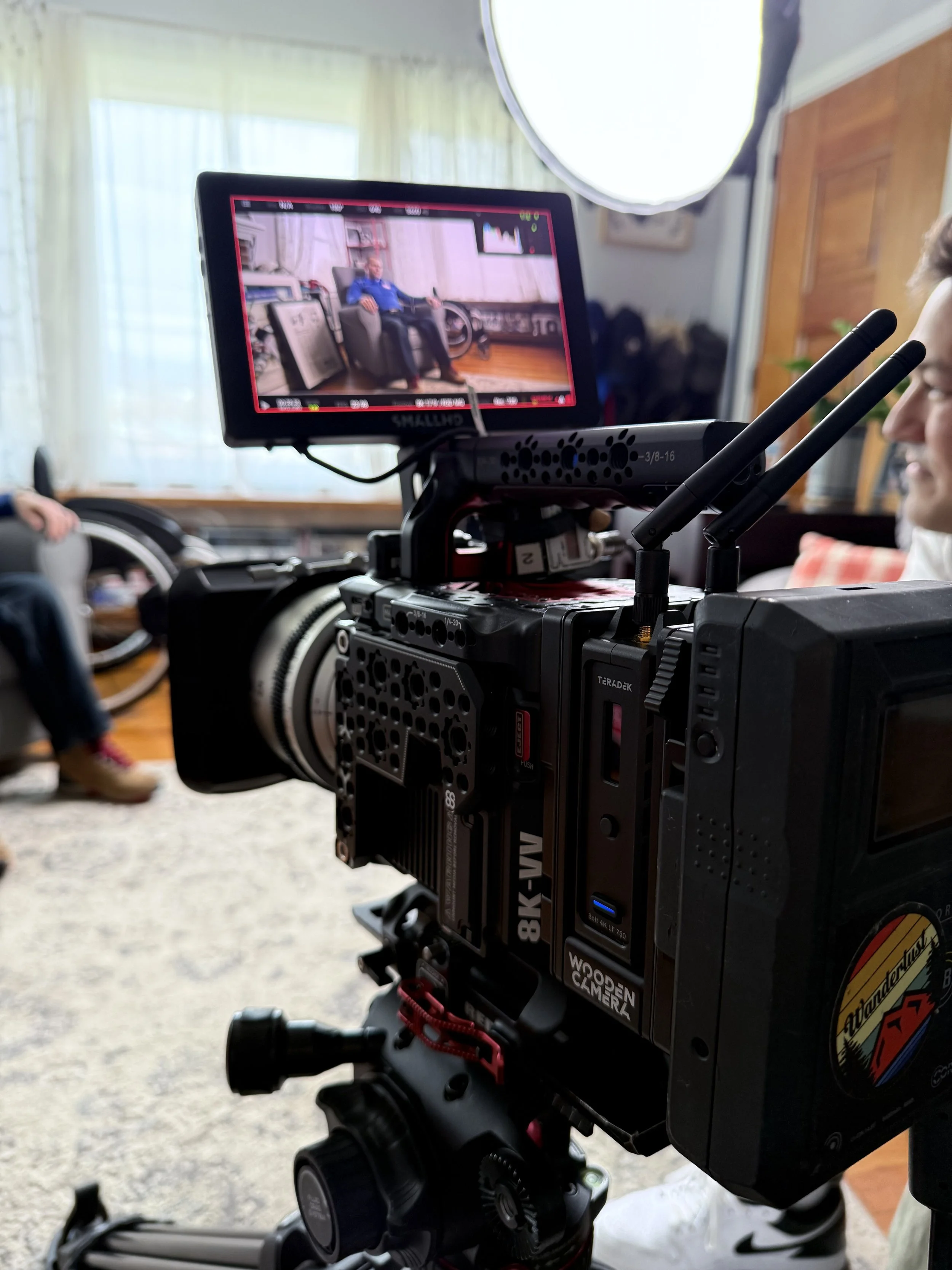 A professional film or photography camera is set up indoors, capturing a scene of a man sitting in a chair in a room with large windows and light-colored curtains. The camera viewfinder screen shows the man sitting in a relaxed pose, wearing a blue s