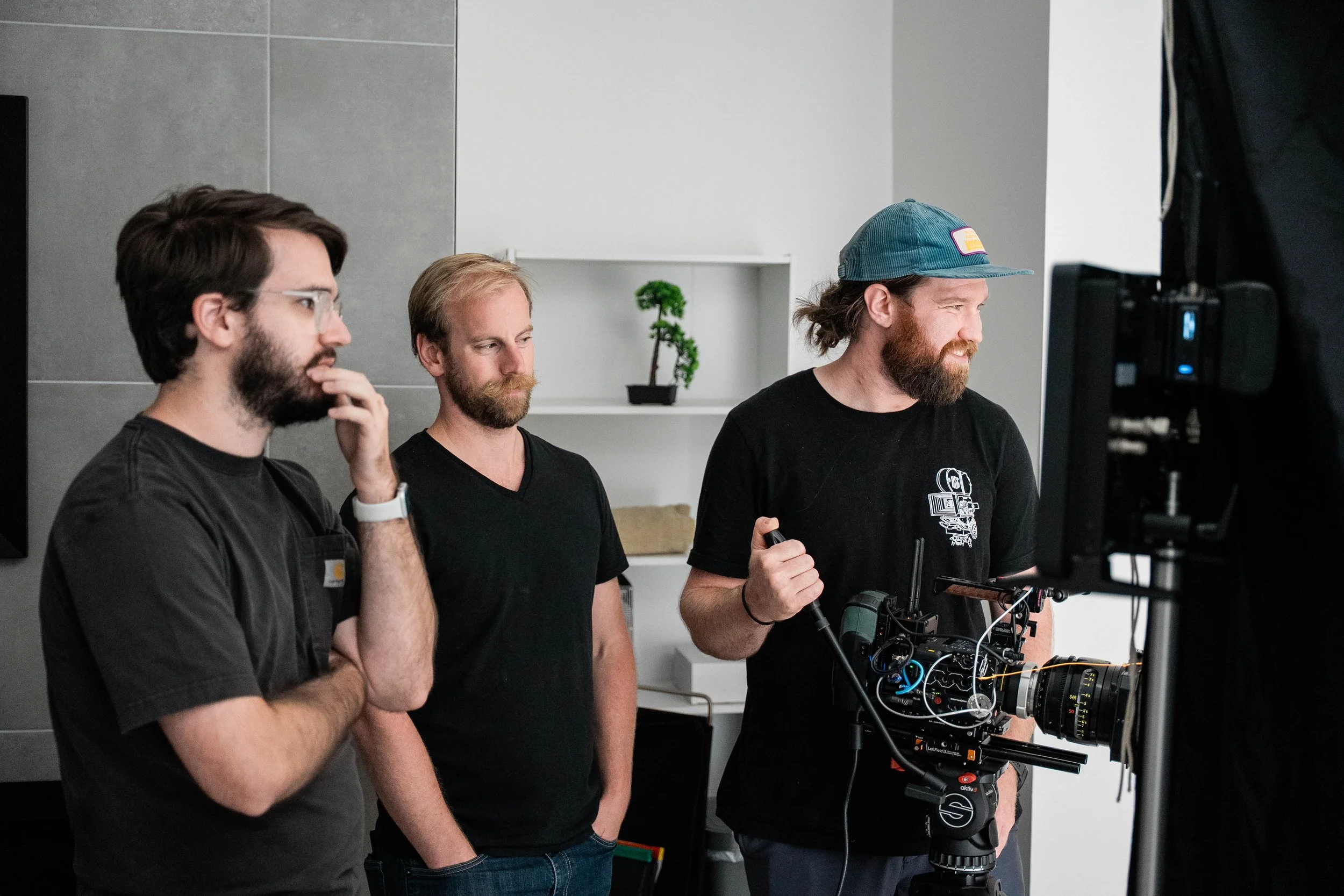 Three men are working together on a film set, with one man operating a professional camera and the others observing and discussing. They are in a modern indoor setting.