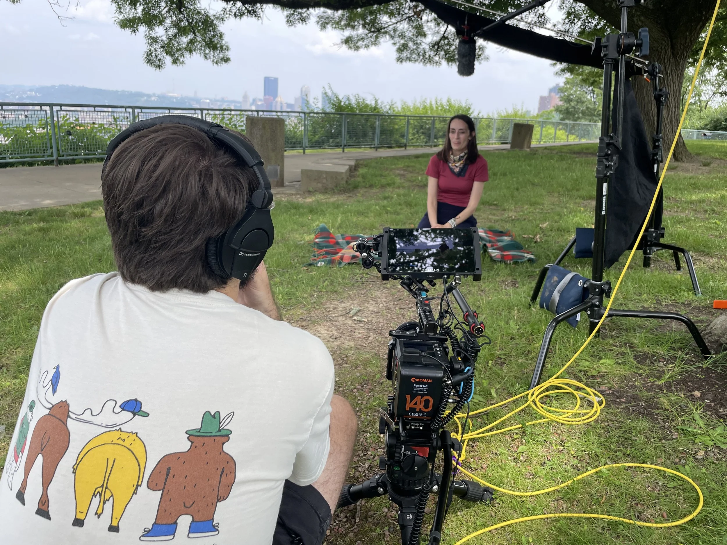 A woman sitting on a blanket outdoors being filmed by a person with professional camera equipment. Another person is operating a camera with headphones. The scene is set in a park with green grass, trees, a city skyline in the background, and cloudy 