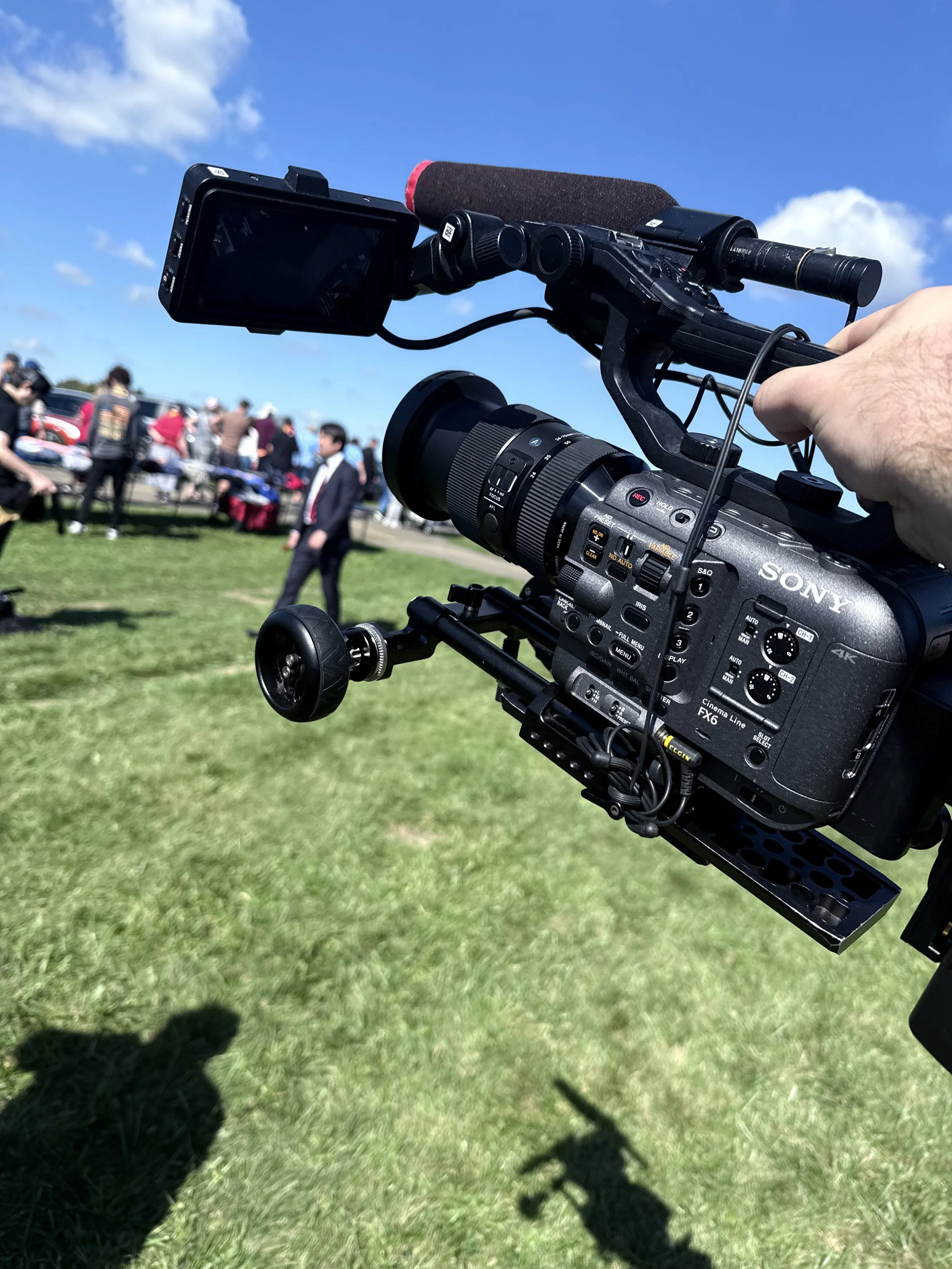 Close-up of a professional video camera mounted on a tripod, focusing on a crowd of people and cars in an outdoor setting on a clear, sunny day with blue sky and scattered clouds.