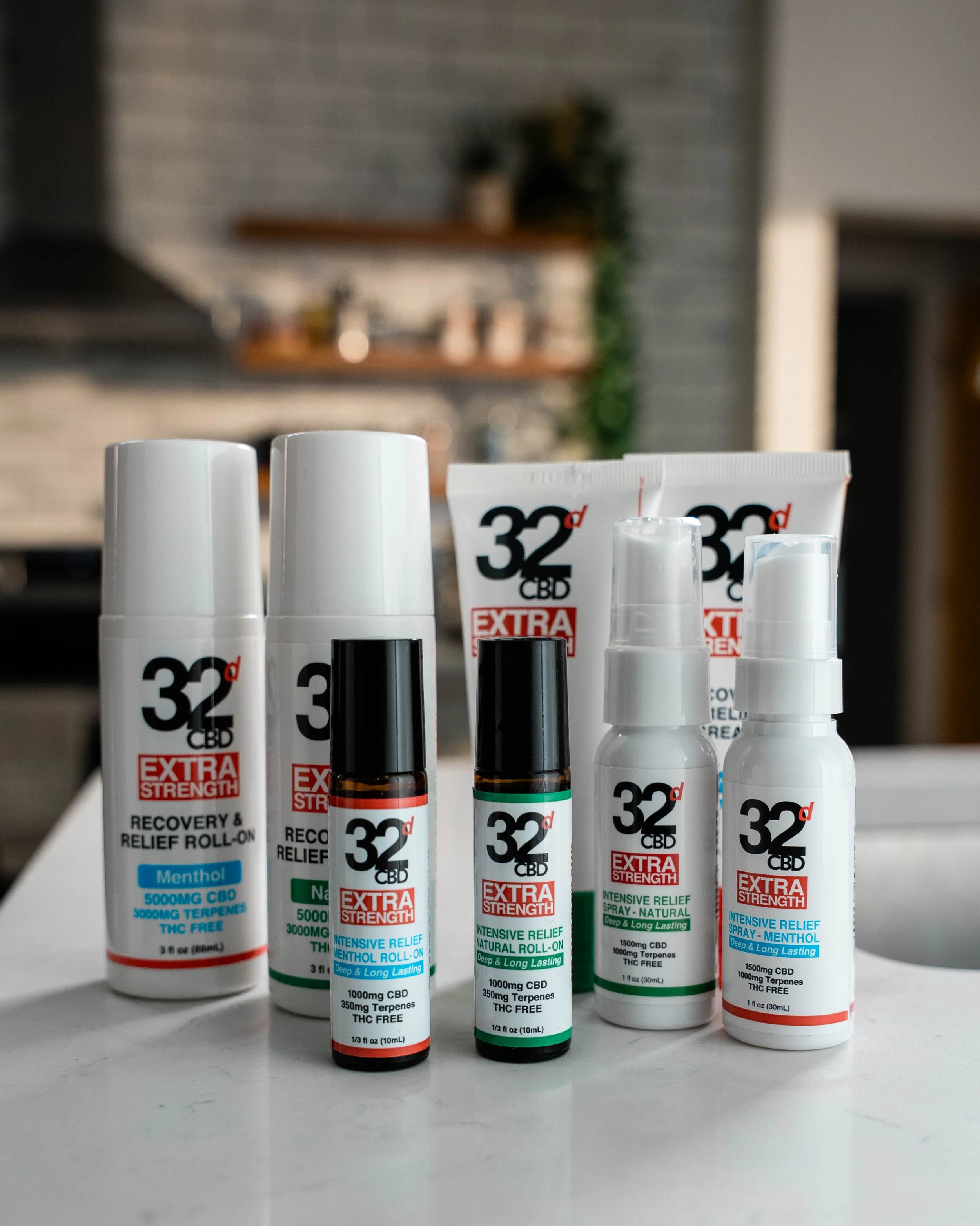 Collection of CBD products including sprays, roll-ons, and creams labeled '32d Extra Strength' on a white surface in a kitchen setting.