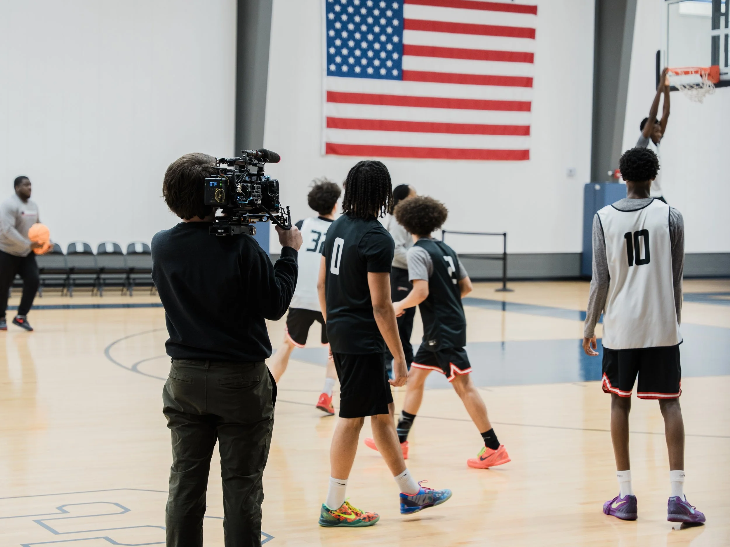A basketball game in progress at an indoor gymnasium, with players on the court and a man filming with a professional camera, and an American flag hanging on the wall.