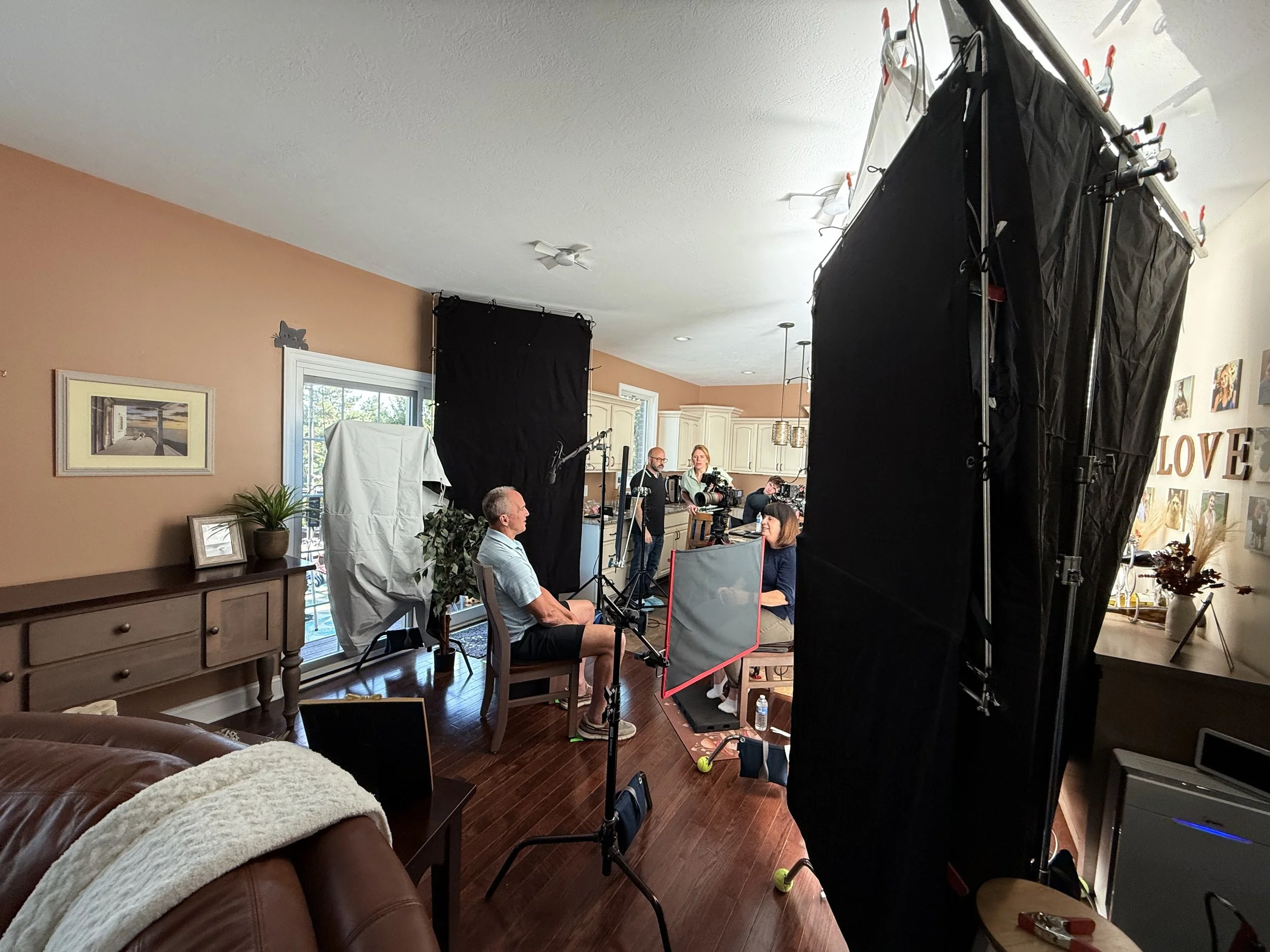 A filming crew is recording a scene inside a house with a peach-colored wall and large windows. Crew members are setting up equipment, and a woman and a man are sitting in front of a black backdrop, possibly being interviewed.