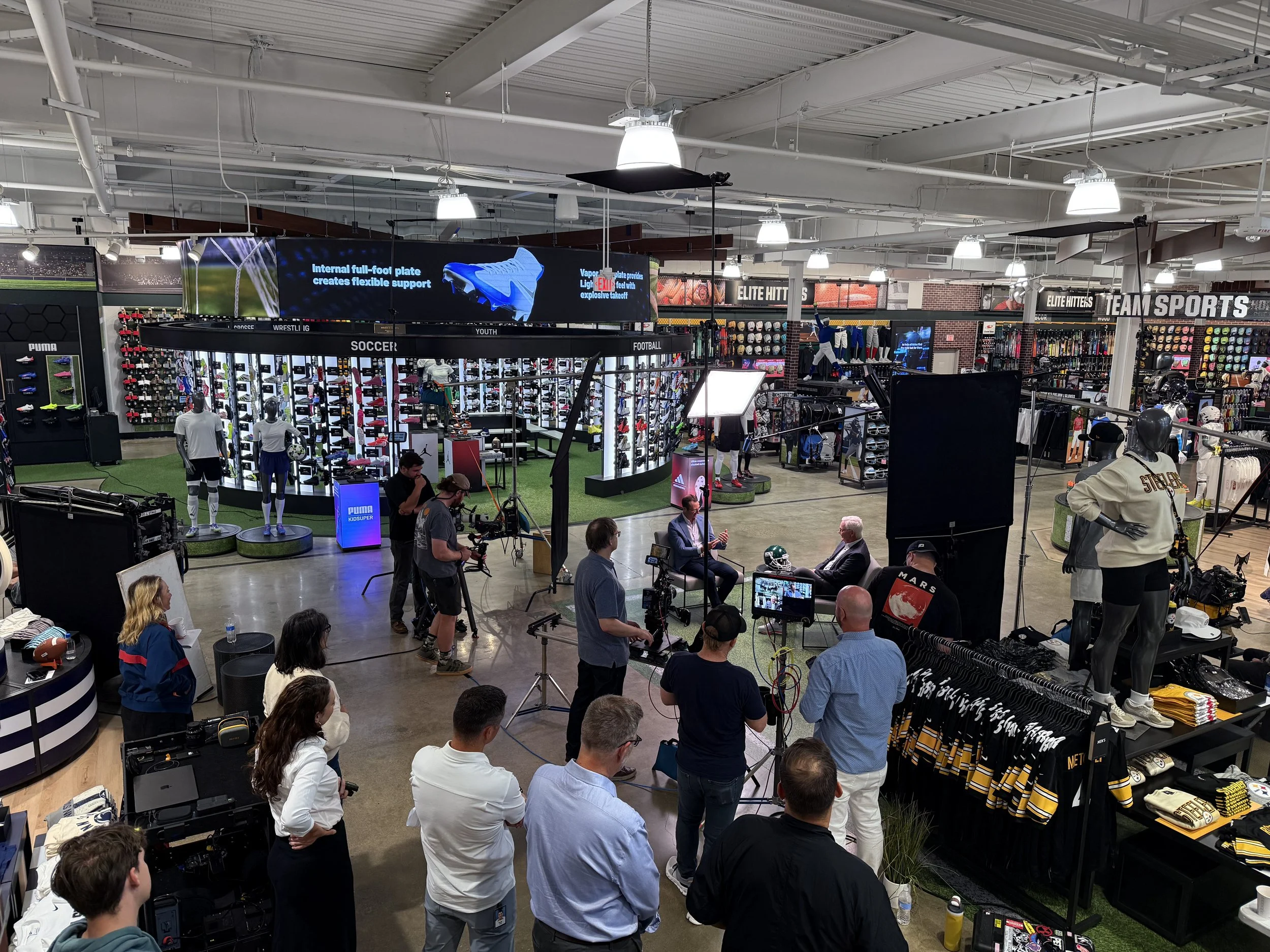 Inside a sports merchandise store with sports apparel, equipment, and mannequins, during a media event or product showcase with people gathering, cameras, and lighting setup.