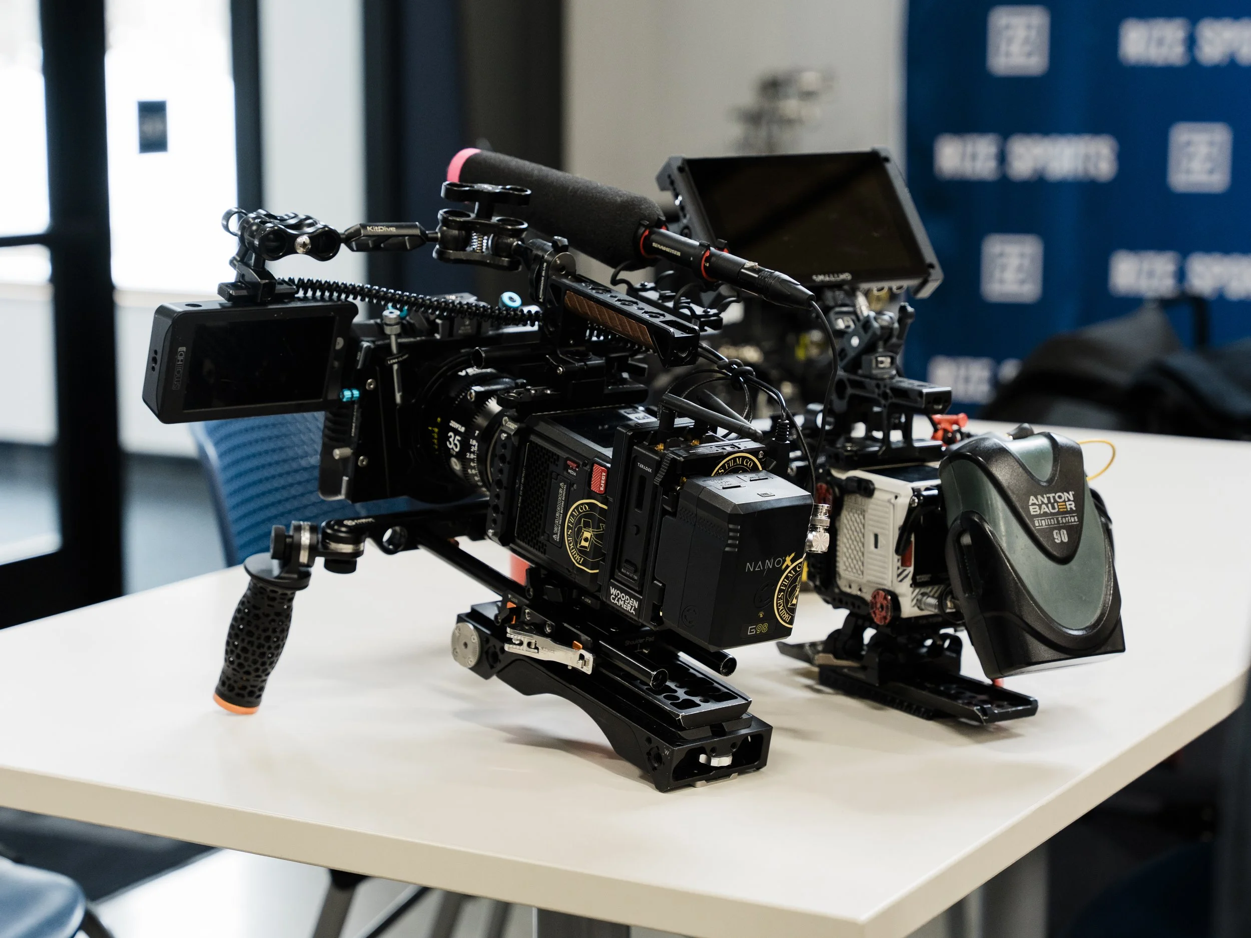 Close-up of professional camera mounted on a stabilizer rig on a white table, with a monitor and microphone attached, in a room with blue chairs and large windows.
