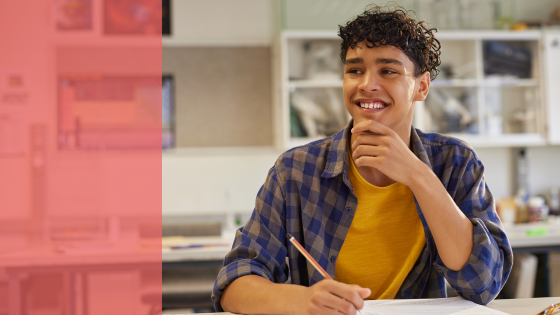 a teen holds a pencil while sitting at a desk. he smiles and looks into the distance