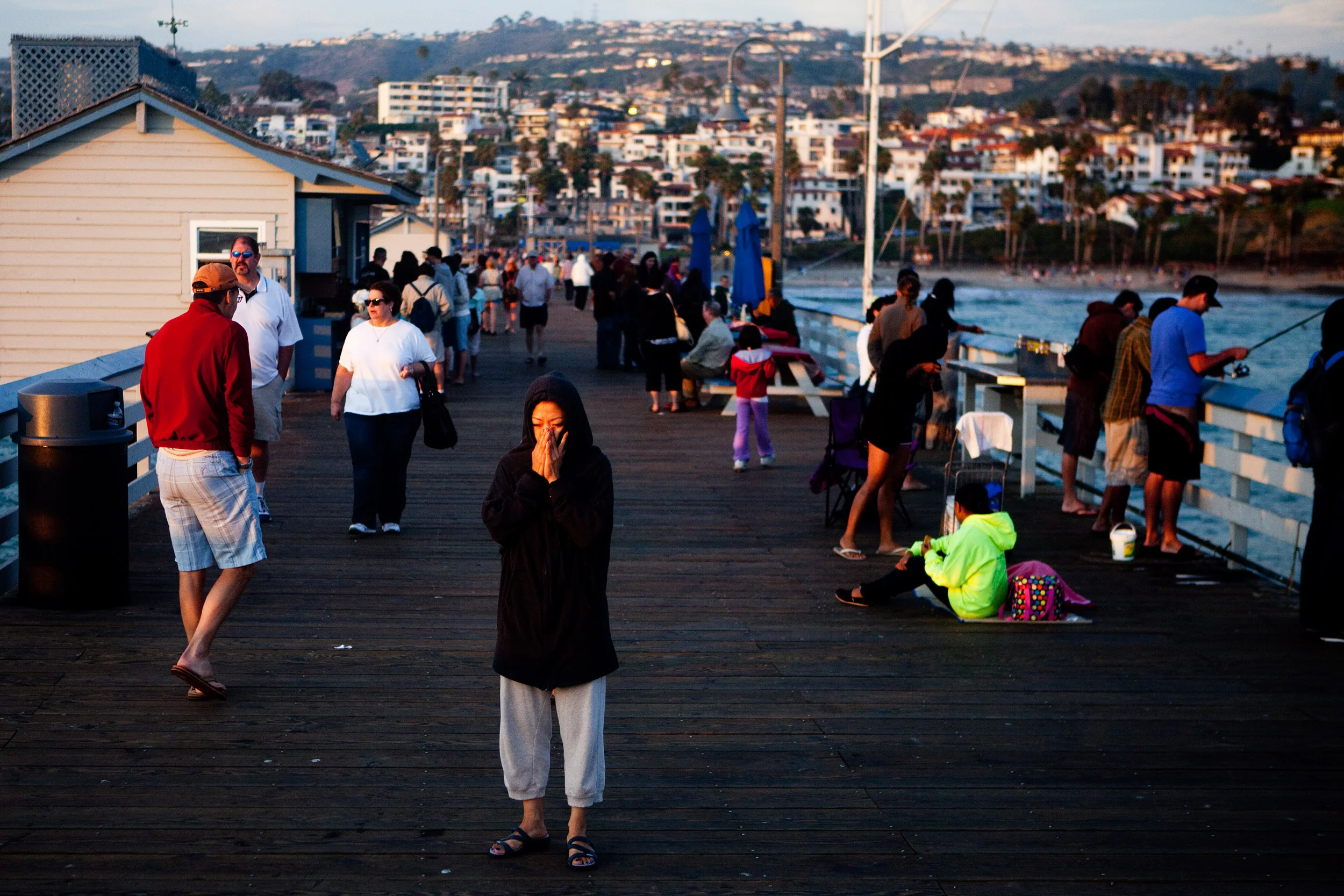 A woman shields her face from the wind while standing near the end of the pier, with a view of the hills of San Clemente, California, behind her.