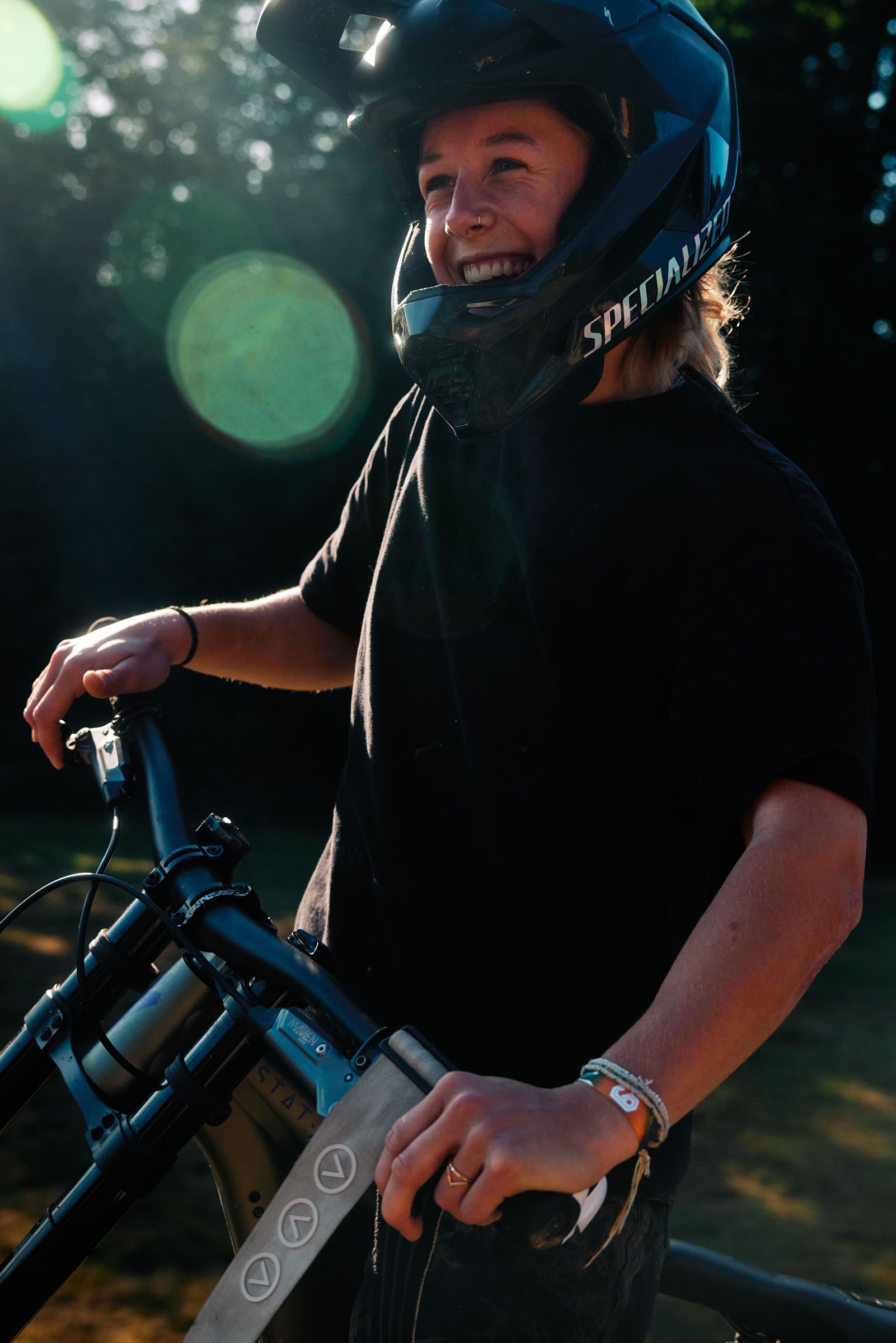 Alma Wiggberg. A person wearing a black helmet with a smile, holding a mountain bike, outdoors in daylight with sun flare in the background.
