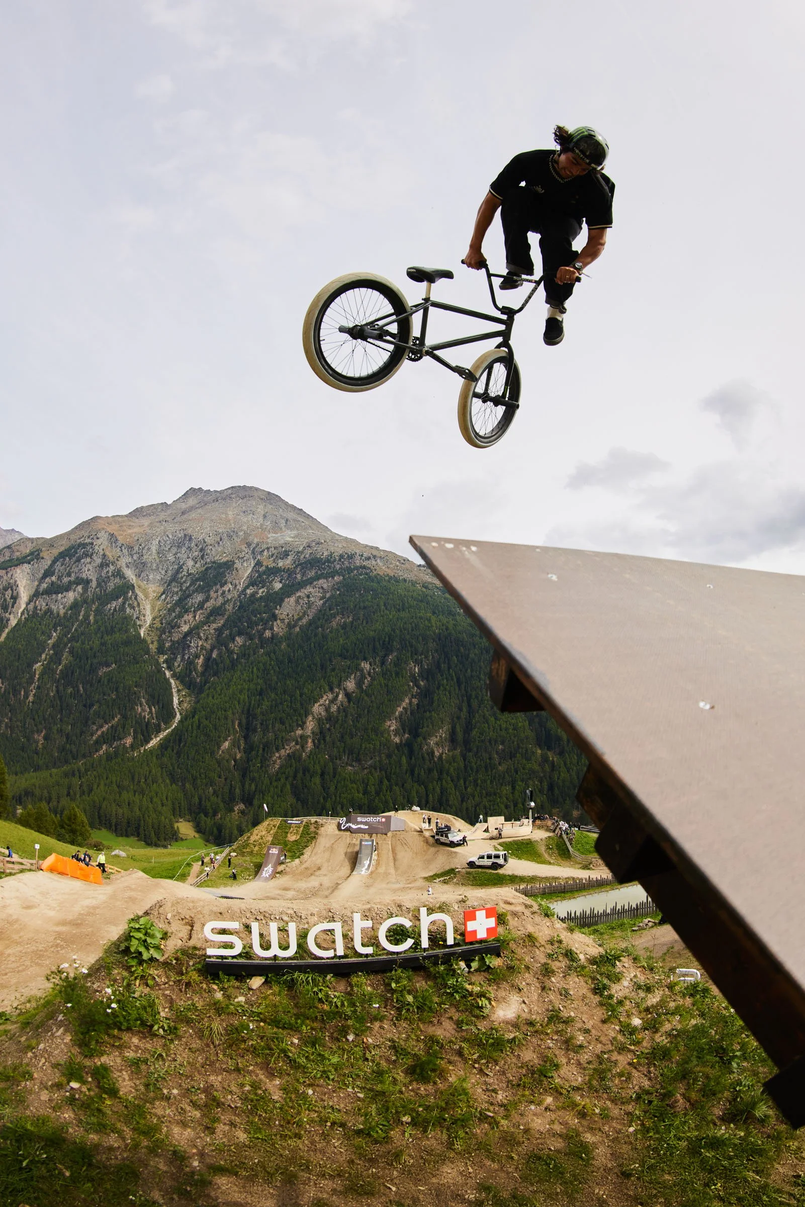 Kevin Peraza Swatch Nines. A BMX rider mid-air performing a trick at a dirt skatepark with a mountain backdrop.