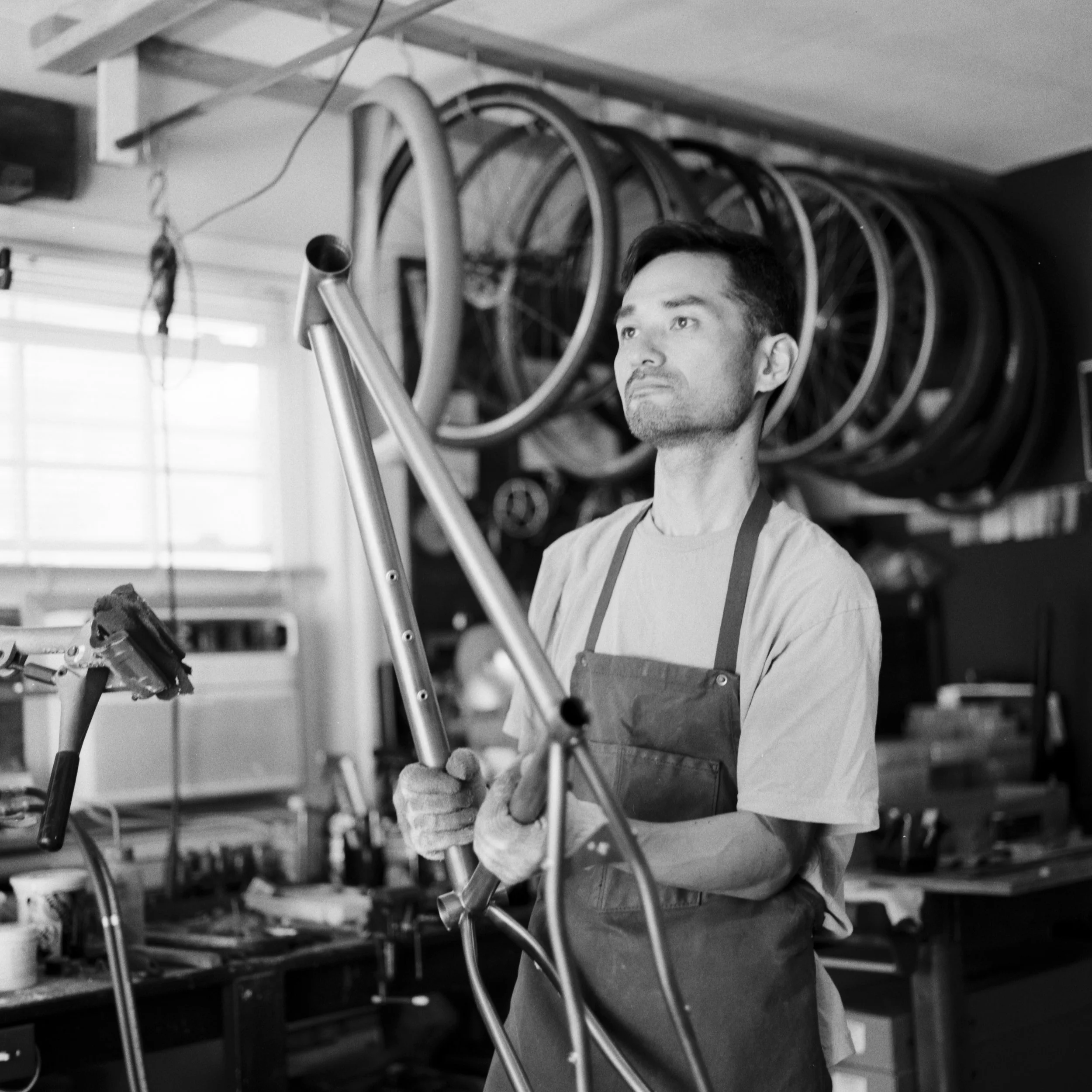 A man working in a bicycle workshop holding a metal bicycle frame, with multiple bicycle wheels hanging on the wall behind him.