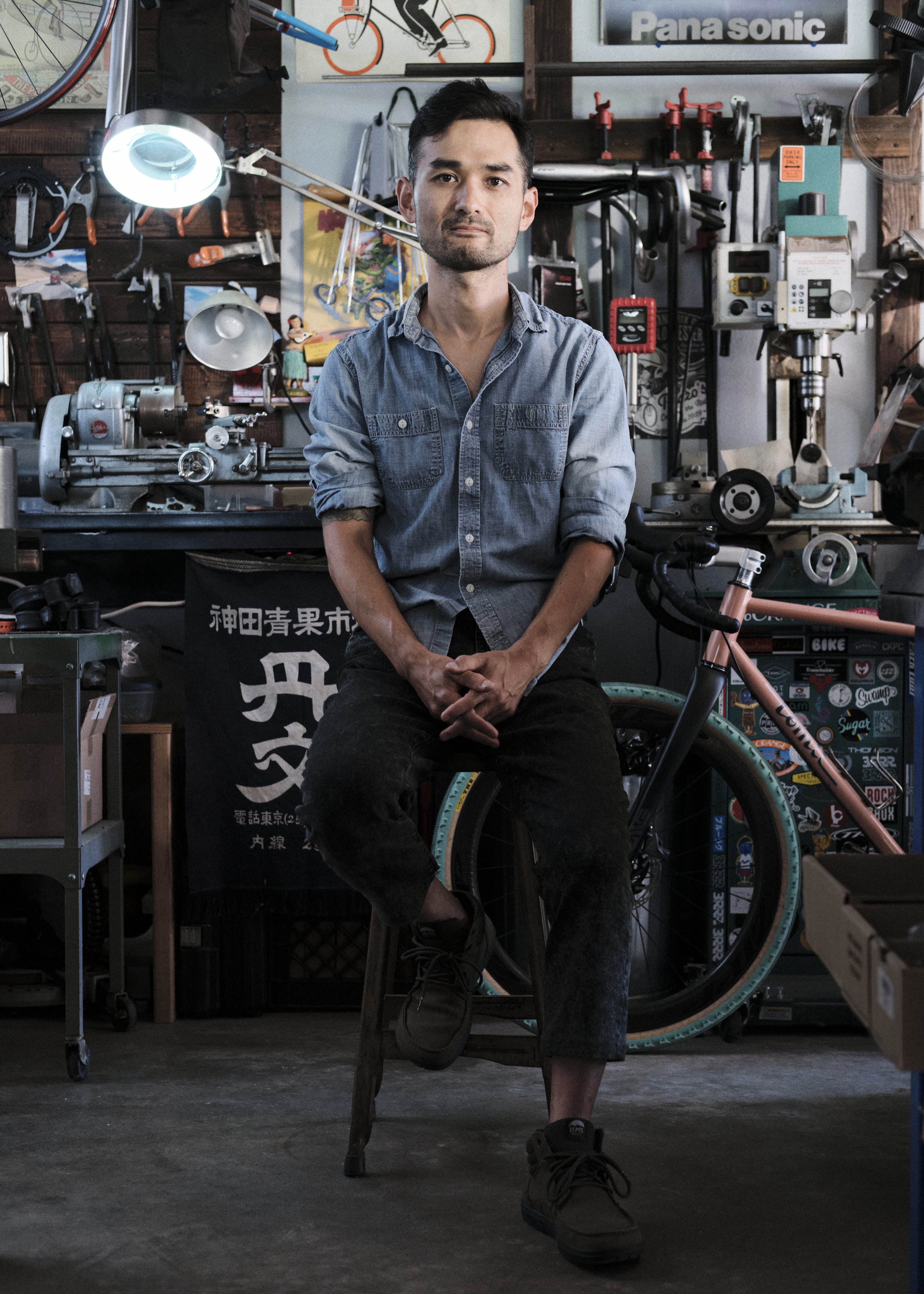 A young man sitting on a wooden stool in a bike workshop, wearing a denim shirt and black pants, with various bicycle parts and tools in the background.