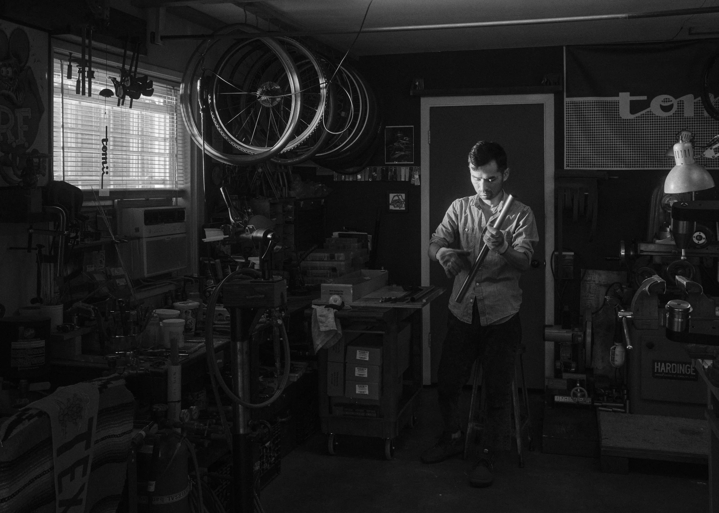 A man working in a cluttered workshop, holding a long metal rod, surrounded by bicycle wheels, tools, and equipment.