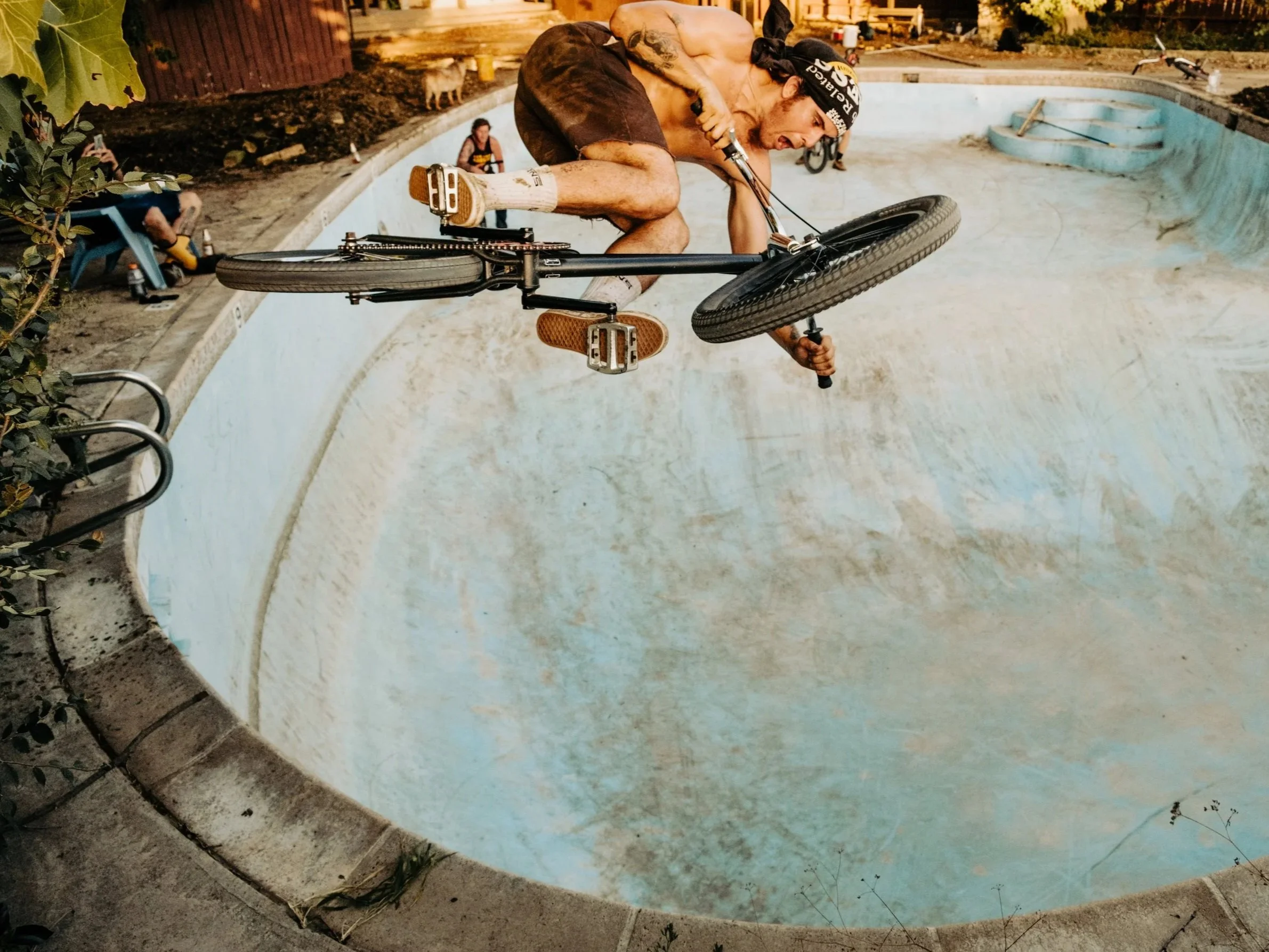 A shirtless man with tattoos riding a BMX bike inside an empty, dirty swimming pool at sunset.
