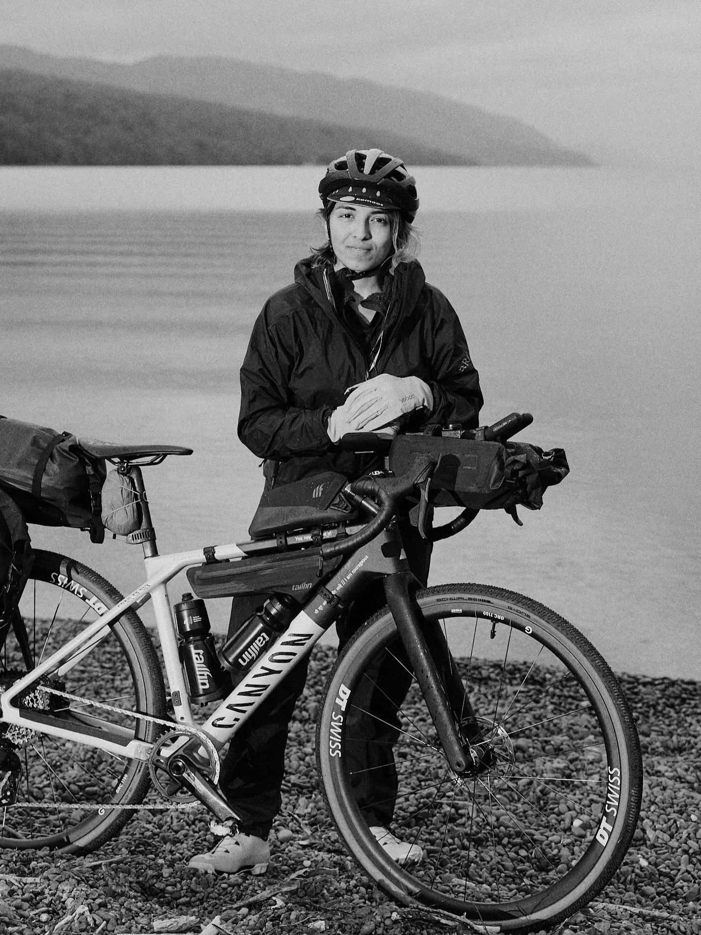 A woman in cycling gear standing with her mountain bike by a lakeside with mountains in the background.