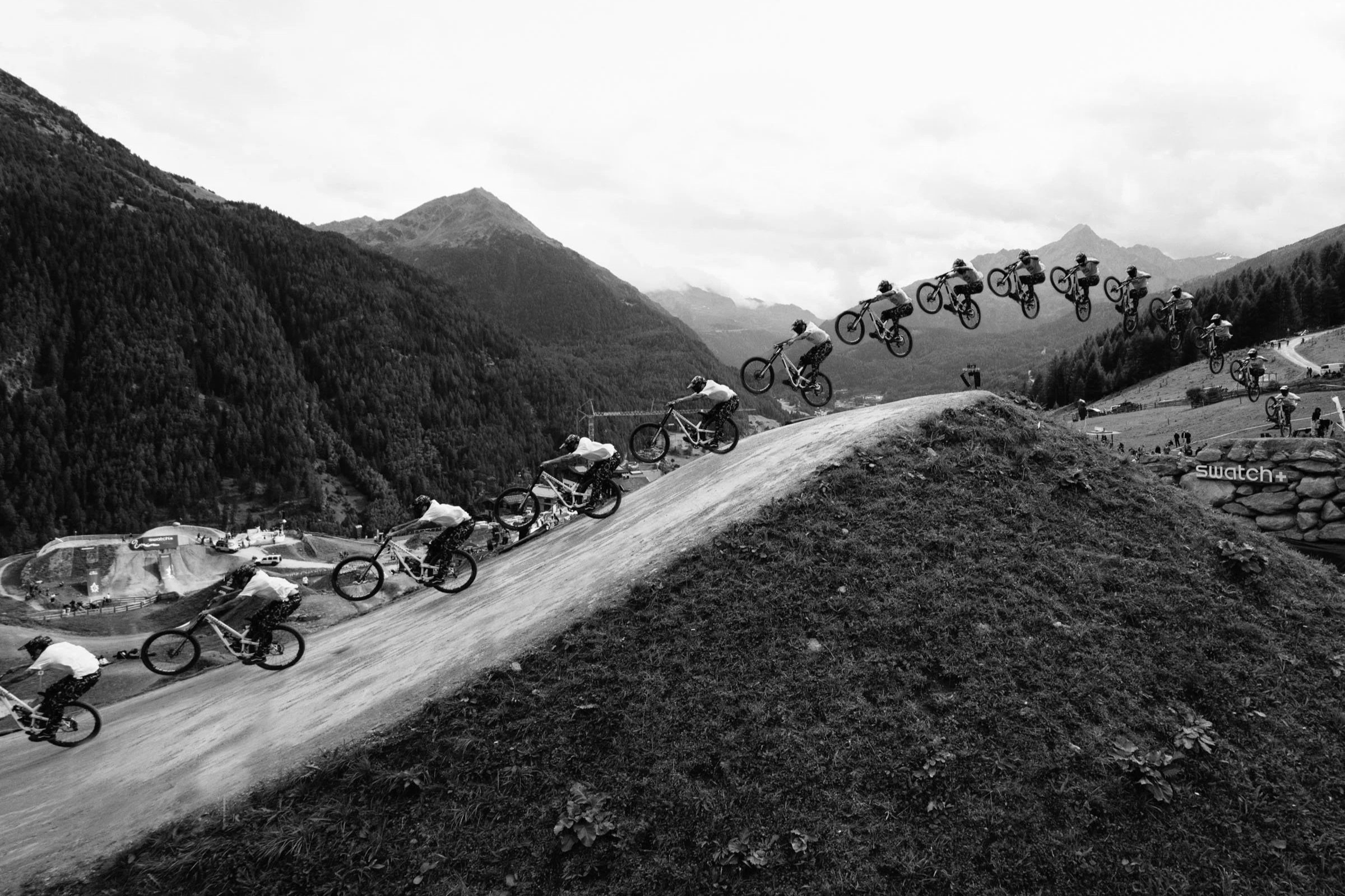 Tomas Lemoine Swatch Nines. Black and white photo of multiple mountain bikers riding down a dirt trail on a hillside with a mountainous landscape in the background.