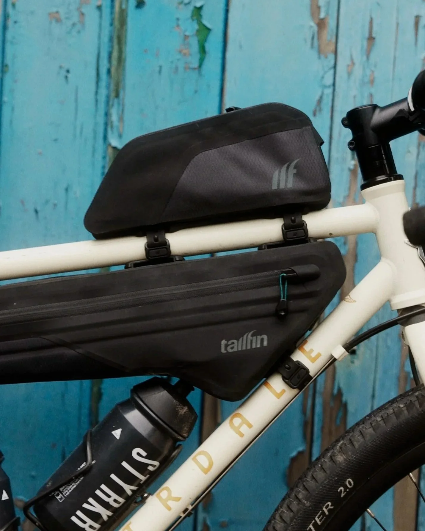 Tailfin Bags. A white bicycle with black storage bags mounted on the frame, against a weathered blue wooden wall.