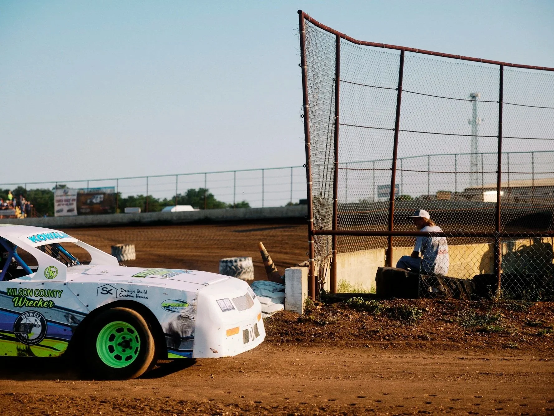 A dirt race track with a partially visible off-white race car with green wheels parked near a rusty chain-link fence. A person wearing a white cap and shirt sits on the ground near the fence, looking at the track. The background shows a clear sky, so