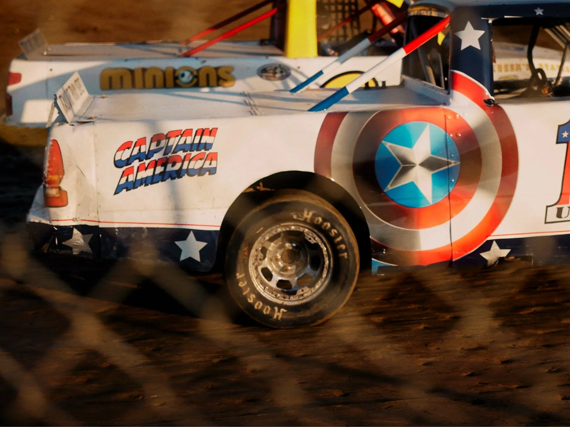 A race car decorated with patriotic American-themed graphics, including a large Captain America shield design, stars, and stripes, racing on a dirt track.
