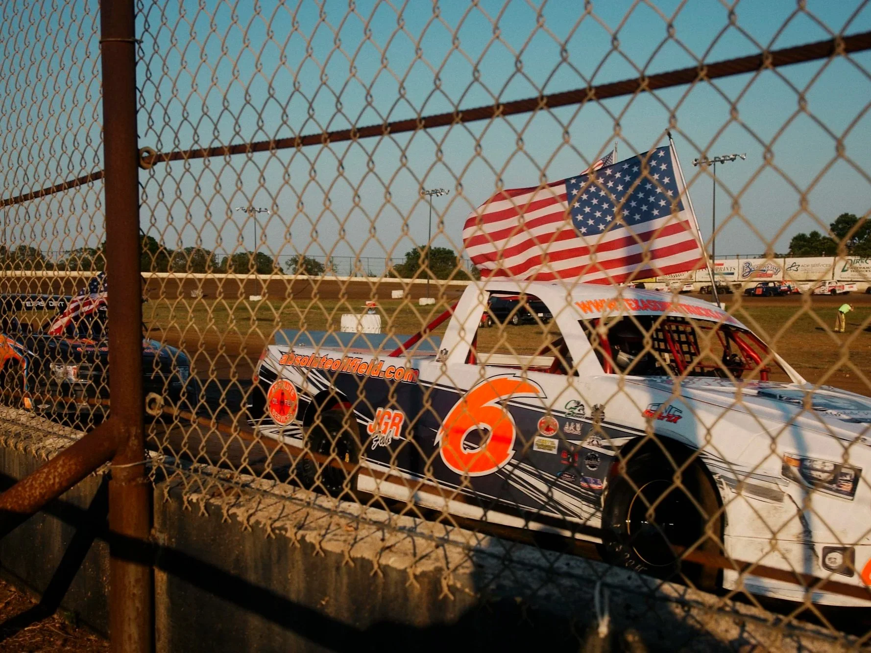 Race cars behind a chain-link fence on a dirt track, with American flags on top of the lead car and in the background during sunset.