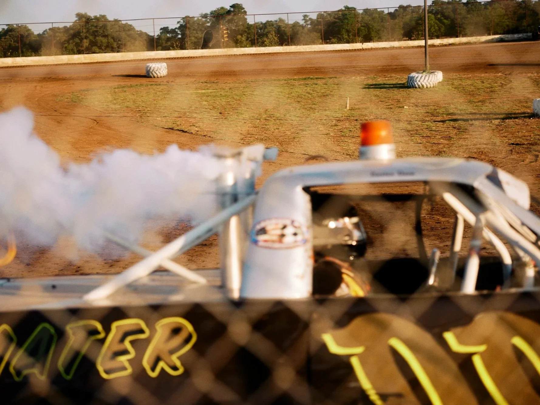 A dirt race track viewed from behind a race car, with dust in the air and tire barriers along the track's edges.