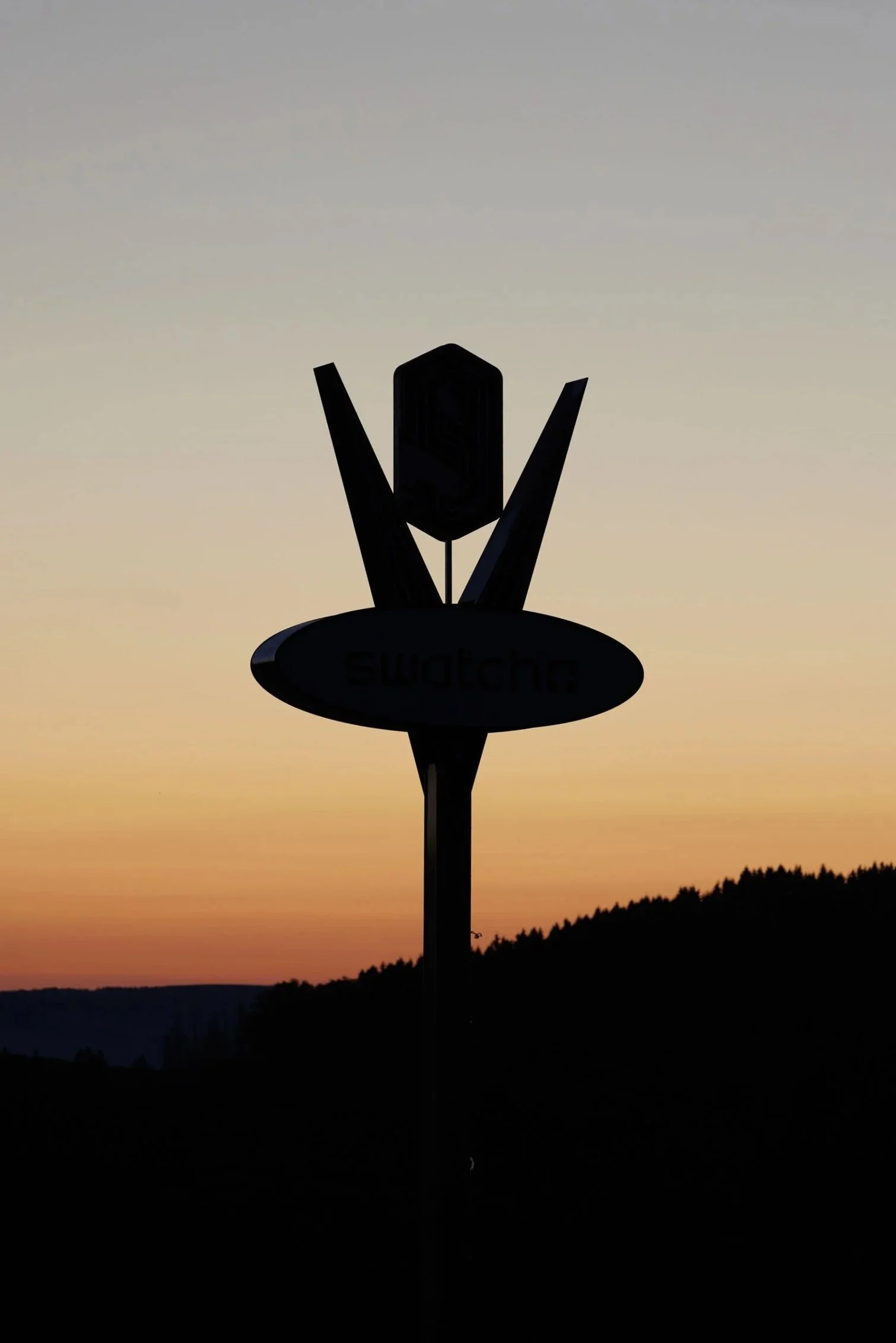 Silhouette of a signpost with the word 'Swatch' against a sunset sky with orange, pink, and purple hues and dark silhouetted mountains.