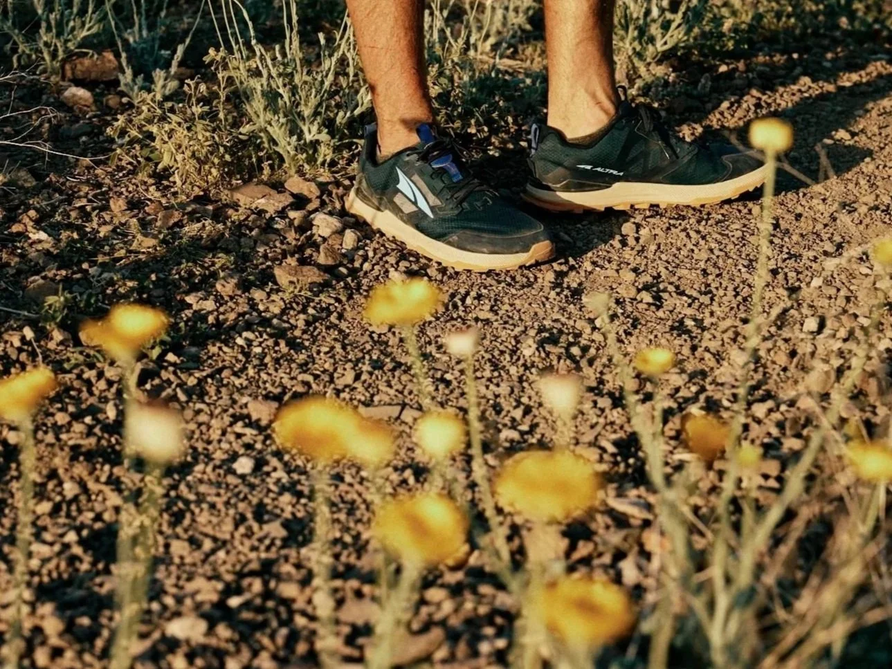 Close-up of a person's legs and feet wearing black trail running shoes, standing on a dirt trail surrounded by small yellow wildflowers and dry grass.