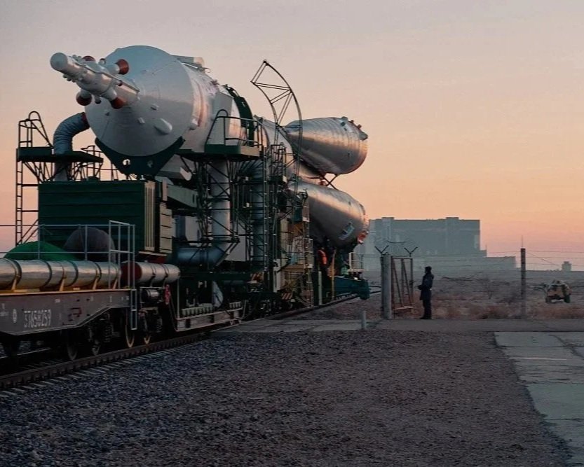 A large rocket being transported on a train in a desert at sunset, with people observing nearby.