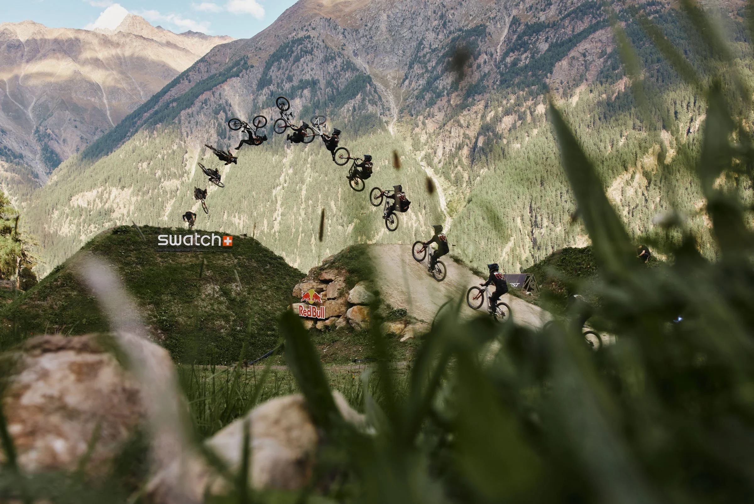 Chance Moore. MTB riders performing jumps and tricks on a dirt track with mountains in the background, surrounded by grass and rocks, featuring Swatch and Red Bull banners.