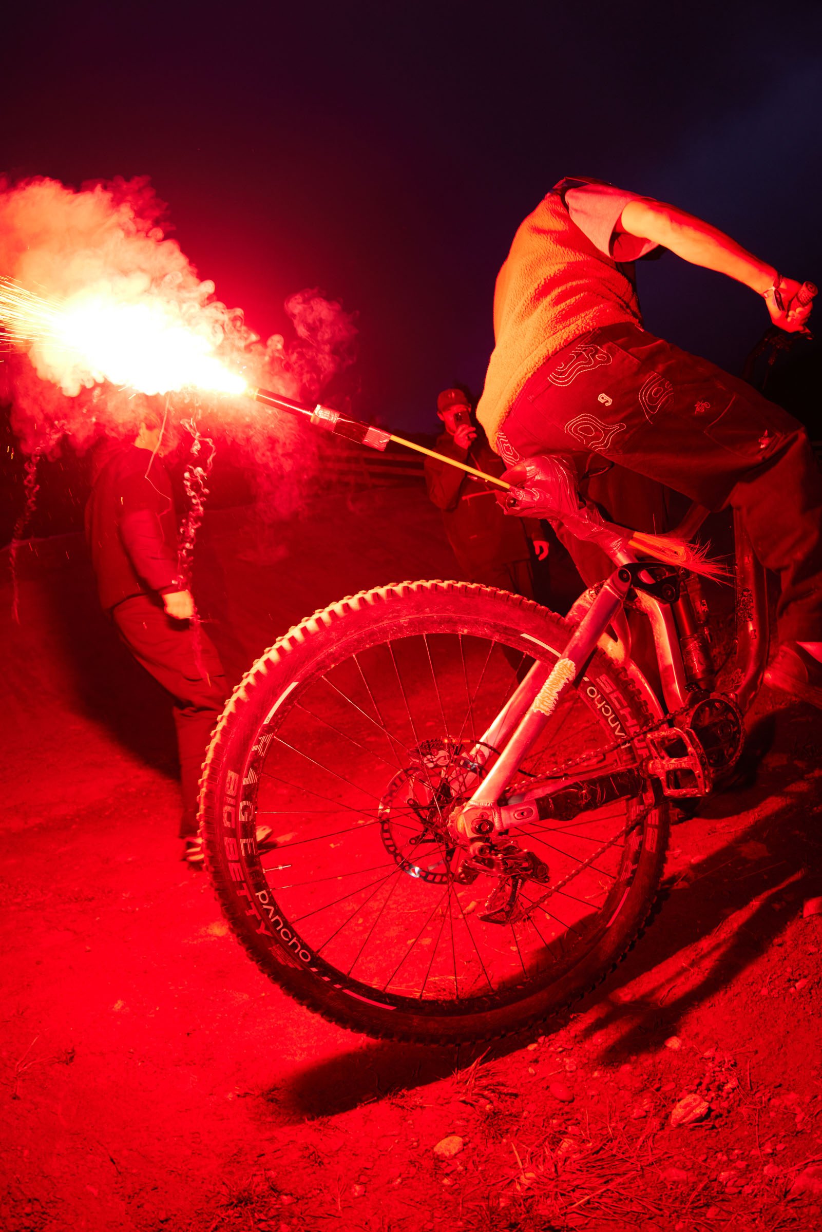 Person riding a mountain bike lit by a red flare at night, with onlookers in the background.