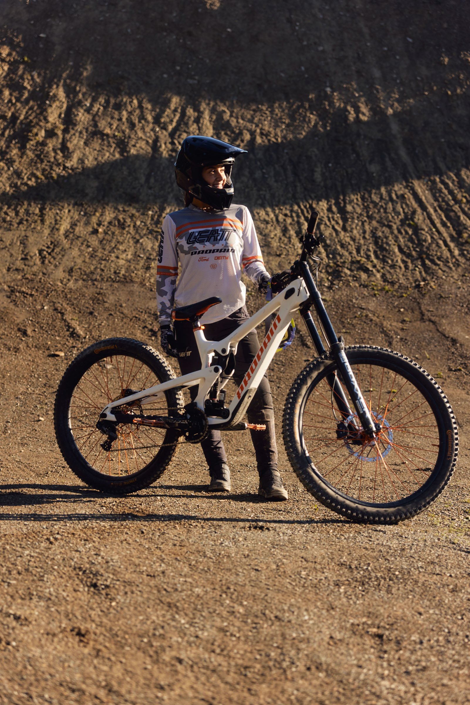 Pascal Sapunar. A woman in mountain biking gear with a helmet, standing beside a white mountain bike on a dirt trail with a dirt hill in the background.