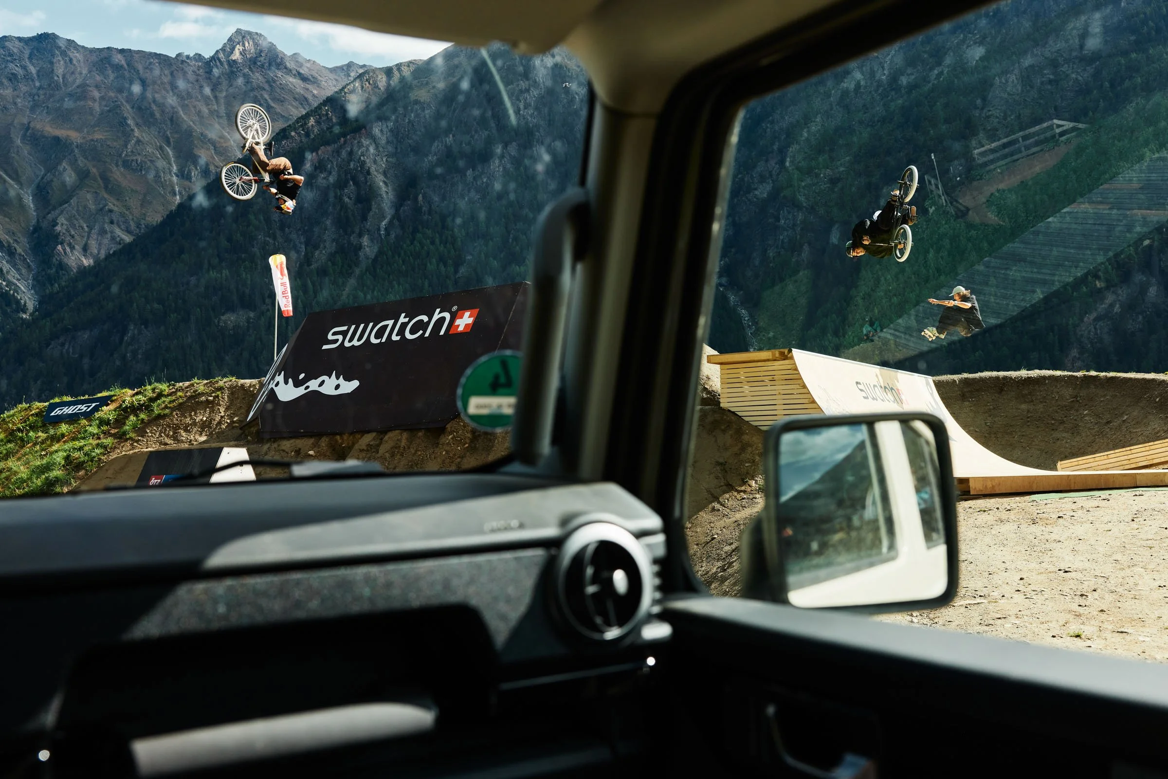 View from inside a vehicle overlooking a mountain dirt skatepark with BMX riders performing tricks in the air against mountain backdrop, Swatch and Red Bull banners visible.