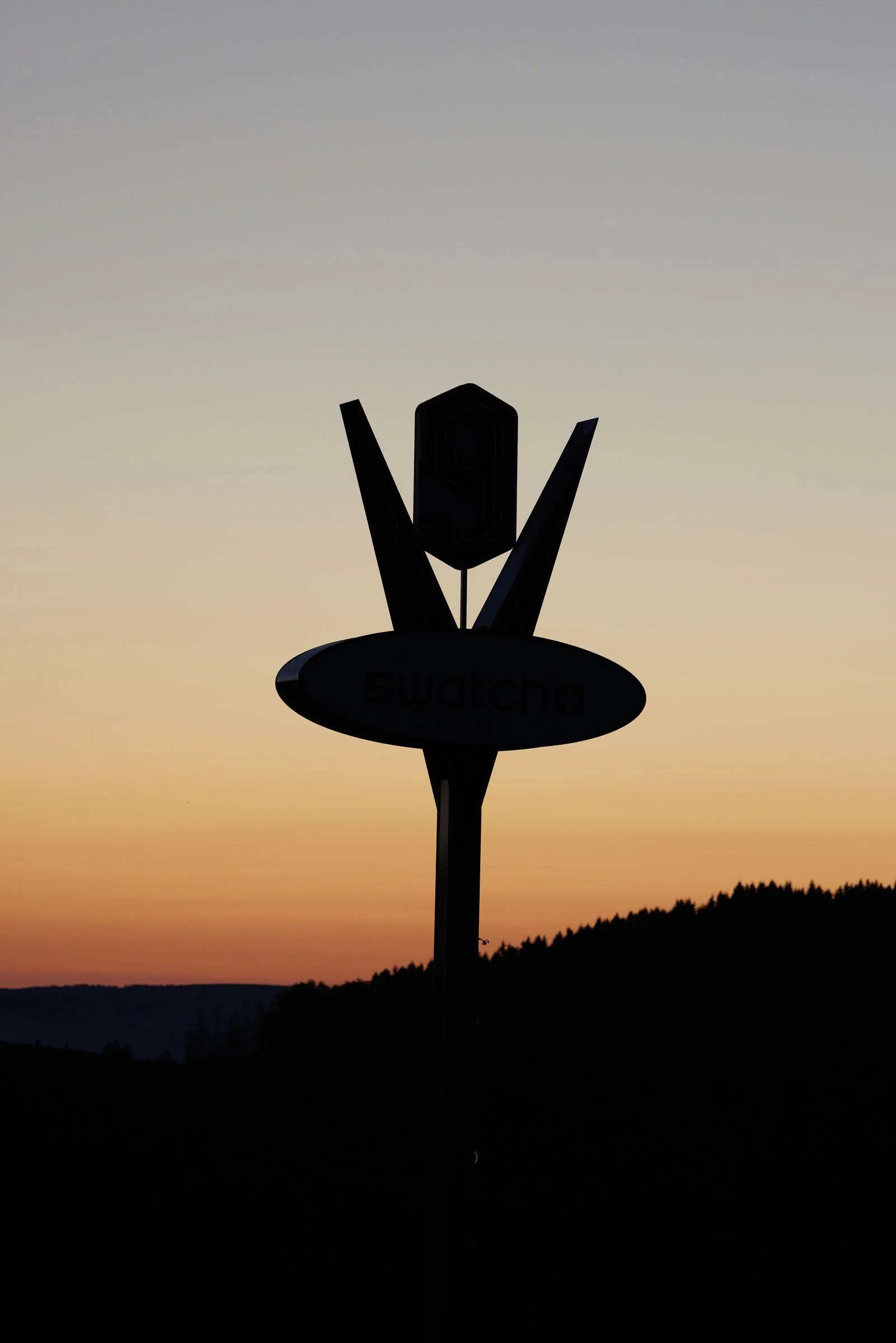 Silhouette of a signpost with the word 'Swatch' against a sunset sky with orange, pink, and purple hues and dark silhouetted mountains.