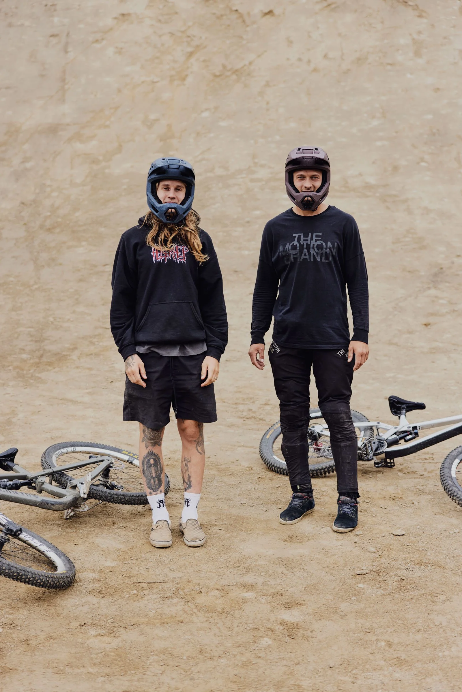 Two mountain bikers wearing helmets and black long-sleeve shirts standing on dirt, with two mountain bikes lying on the ground beside them, against a beige dirt wall.
