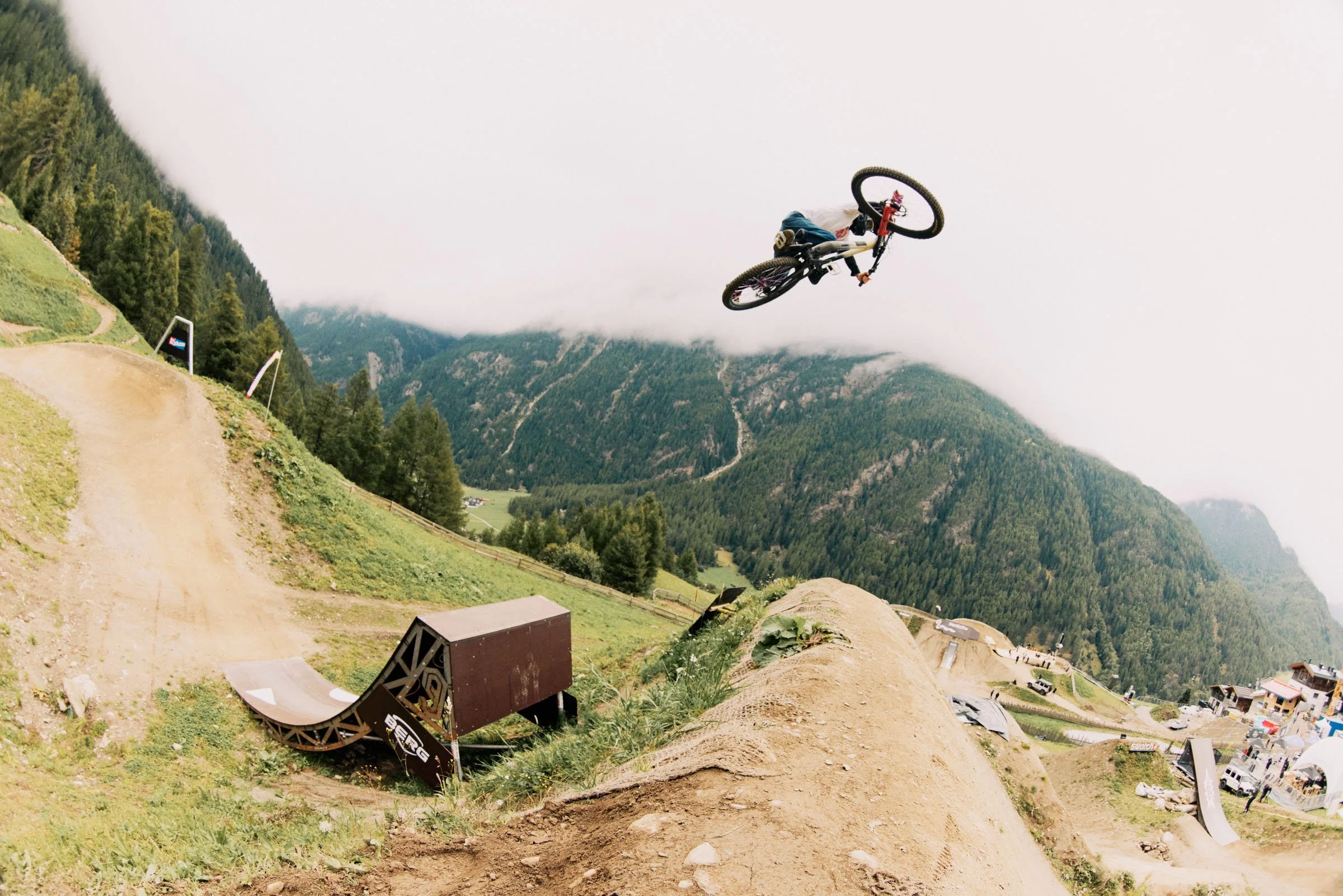 A mountain biker performing a jump off a dirt ramp on a dirt track in a mountainous area with green hills and trees, under a cloudy sky.