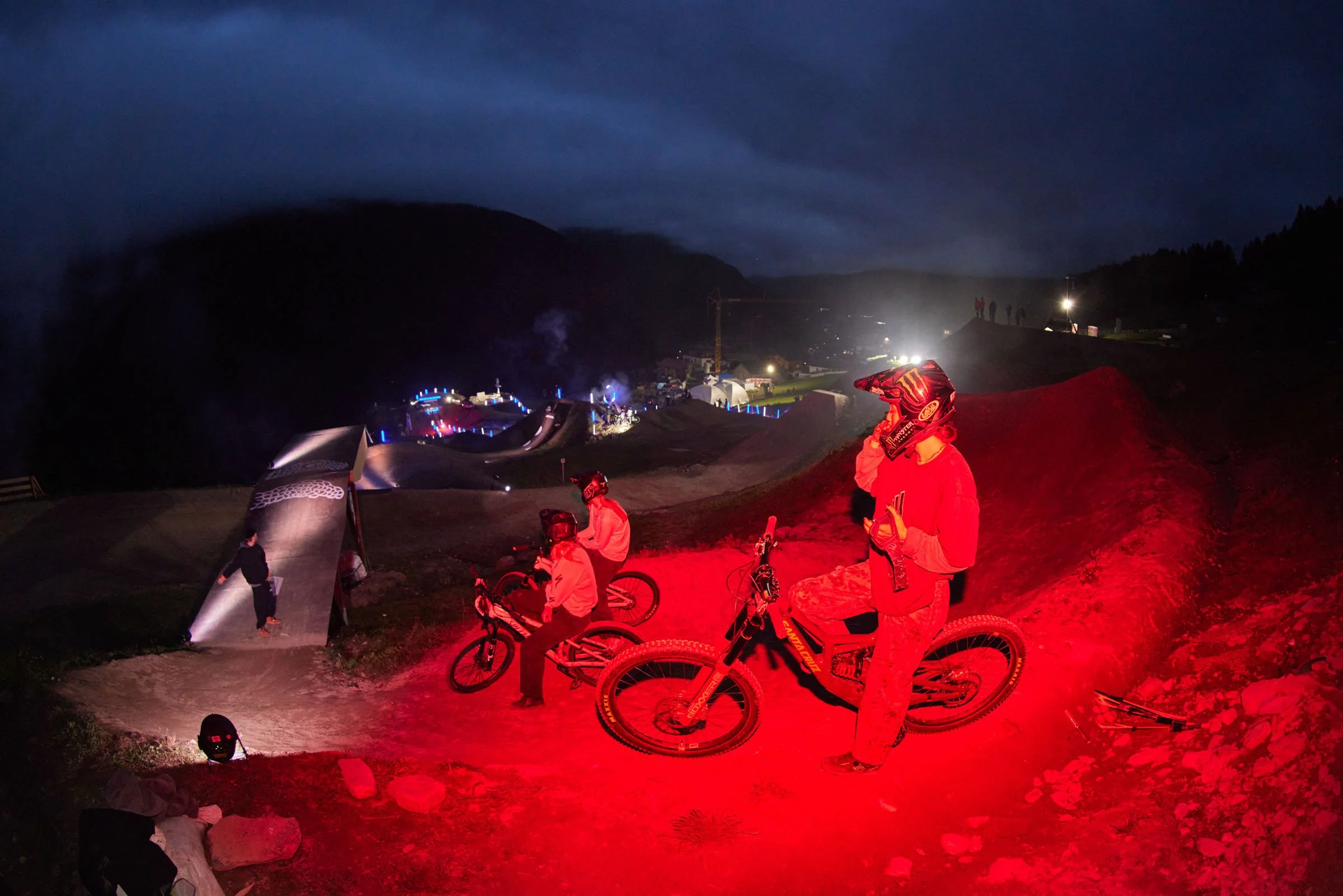 Nighttime mountain biking scene with three cyclists in red clothing and helmets resting on their bikes, illuminated by red light, with a BMX track in the background, and a dark mountain landscape under a cloudy sky.
