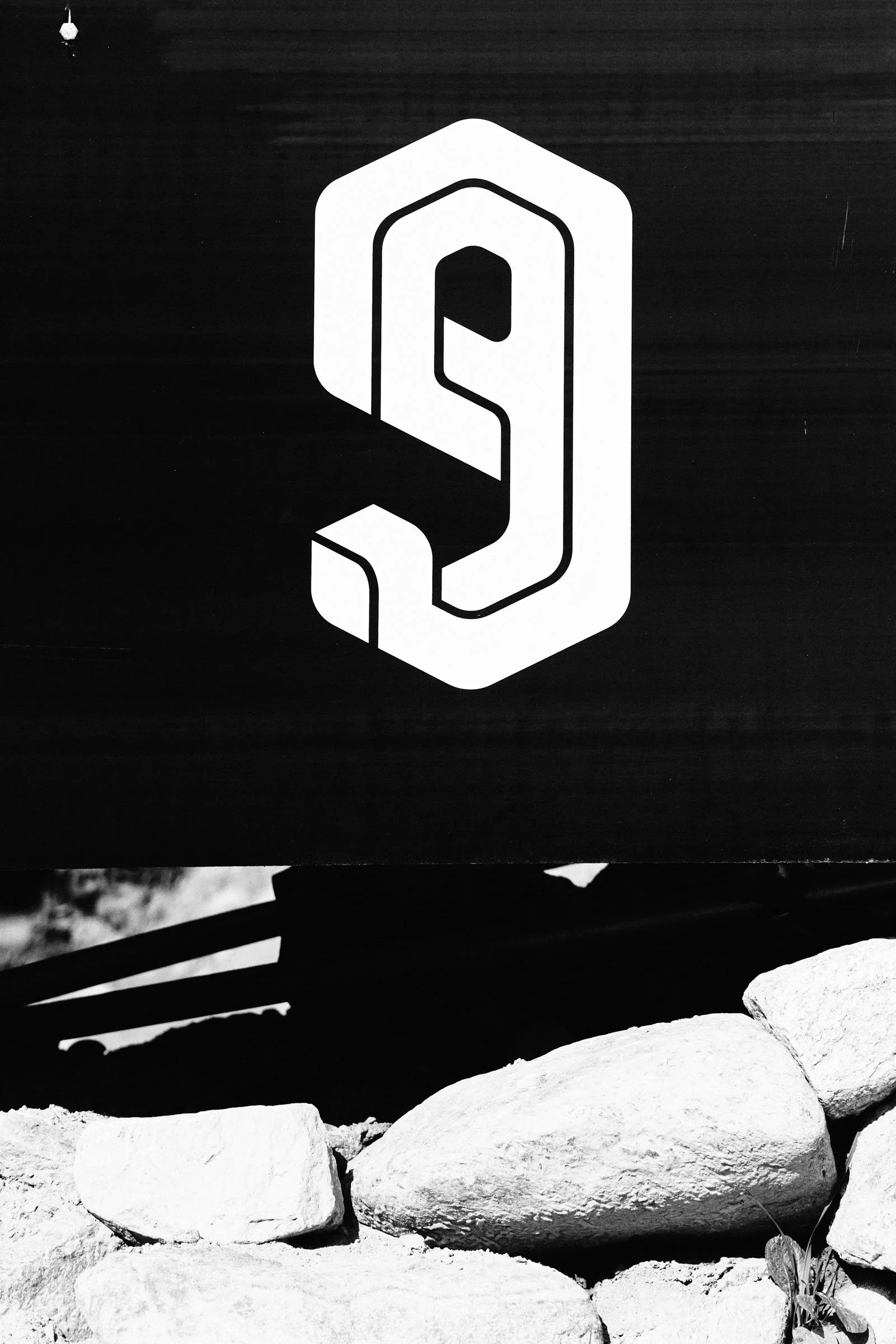 Black and white image of a sign with a stylized number nine or letter 'P' on a dark background, placed above a pile of rocks.
