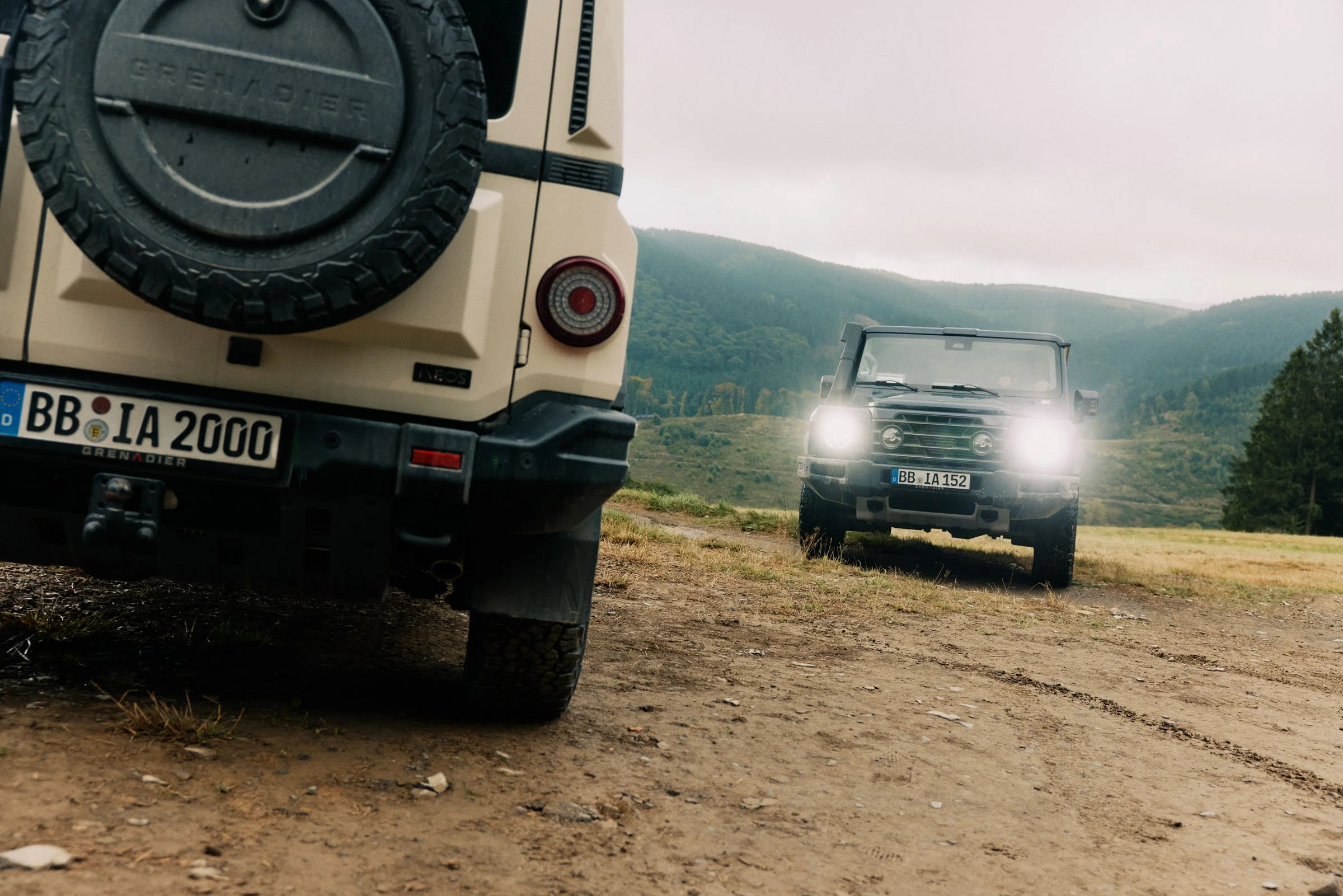 Two off-road vehicles on a dirt path in a rural area with green hills and trees in the background. The vehicle in the foreground is a beige off-road vehicle with a spare tire mounted on the back, and the vehicle in the distance has its headlights on.