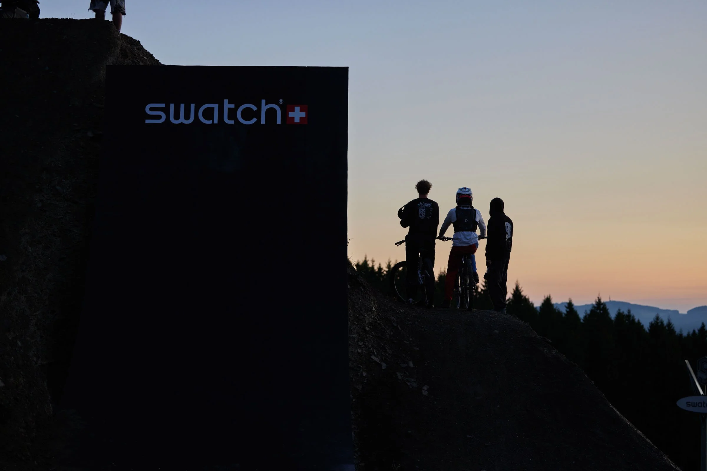 Three people, one with a bicycle, standing on a hill at sunset near a Swatch sign.