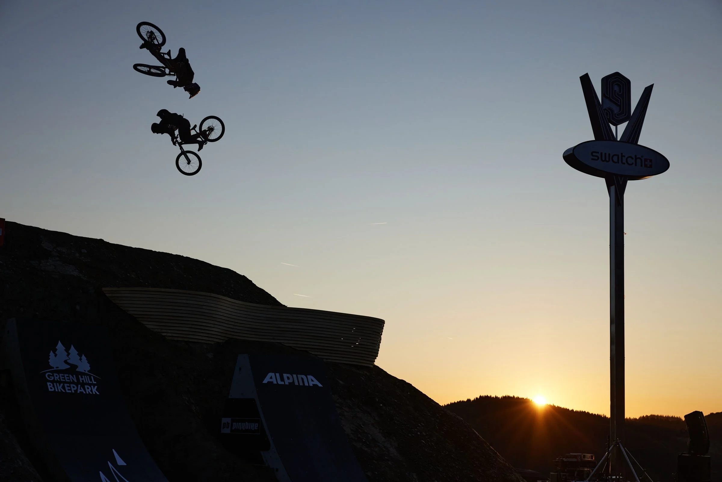 Szymon Godziek. Two BMX riders performing tricks in mid-air during sunset at Green Hill Bikepark, with a large Swatch sign and mountainous landscape in the background.