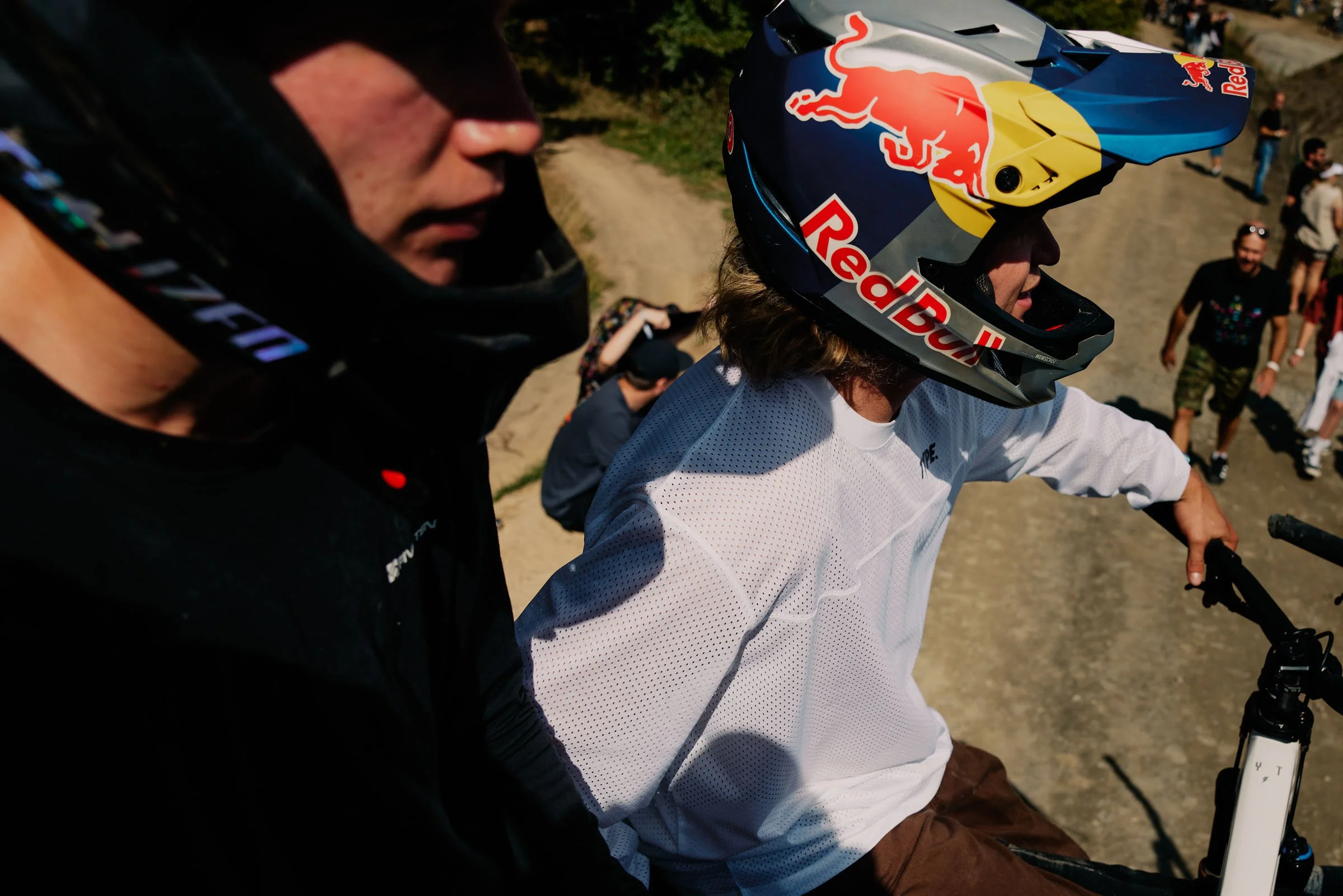 Erik Fedko, Chance Moore. Two cyclists wearing helmets and outdoor gear, one with a Red Bull helmet, on a dirt trail with people in the background.